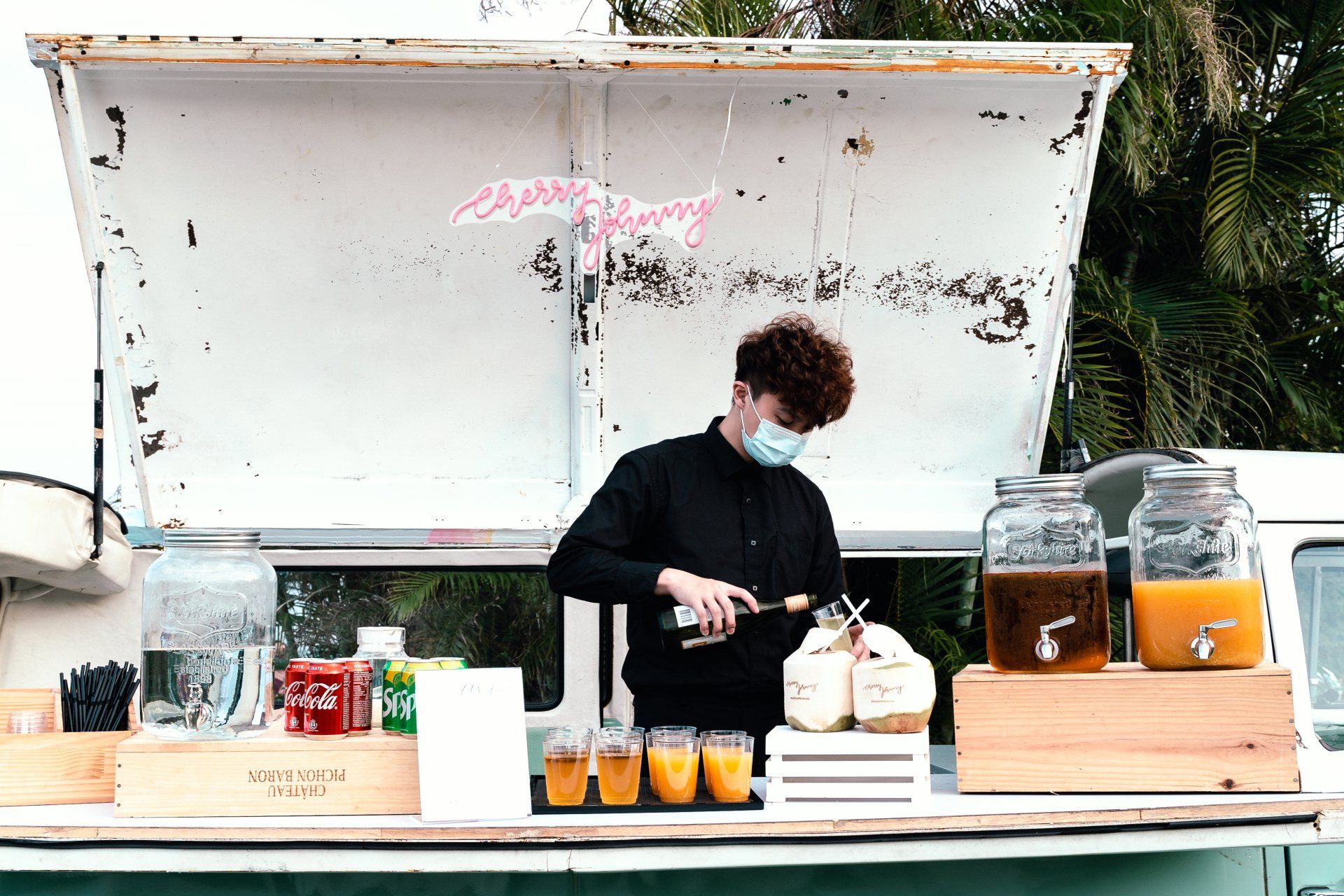 Man in mask pours liquid into coconut at a drink stand with various beverages.