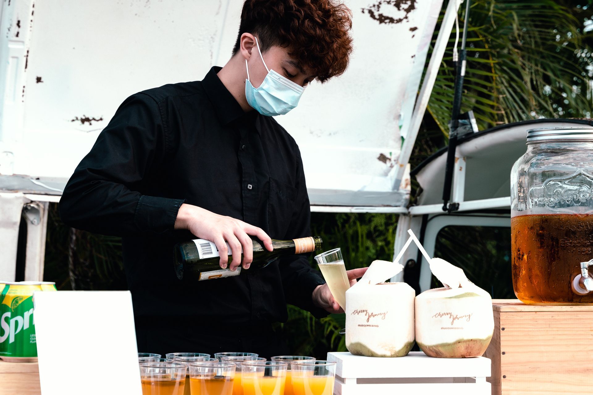 Bartender in mask pouring drink from a bottle into a glass, with coconut drinks nearby.