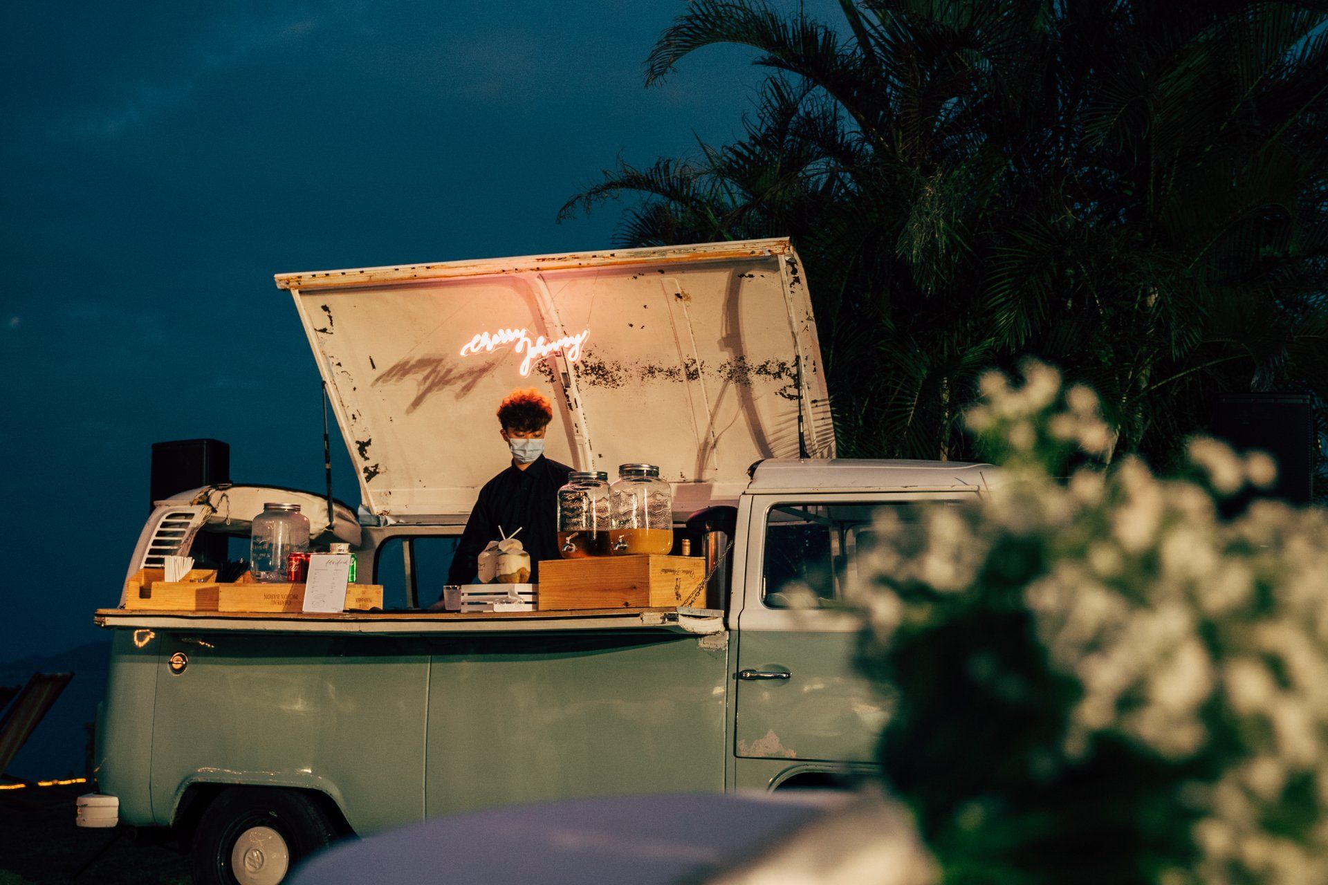 Food truck with open hatch, barista preparing drinks. Blue-green exterior, neon sign, dark background.