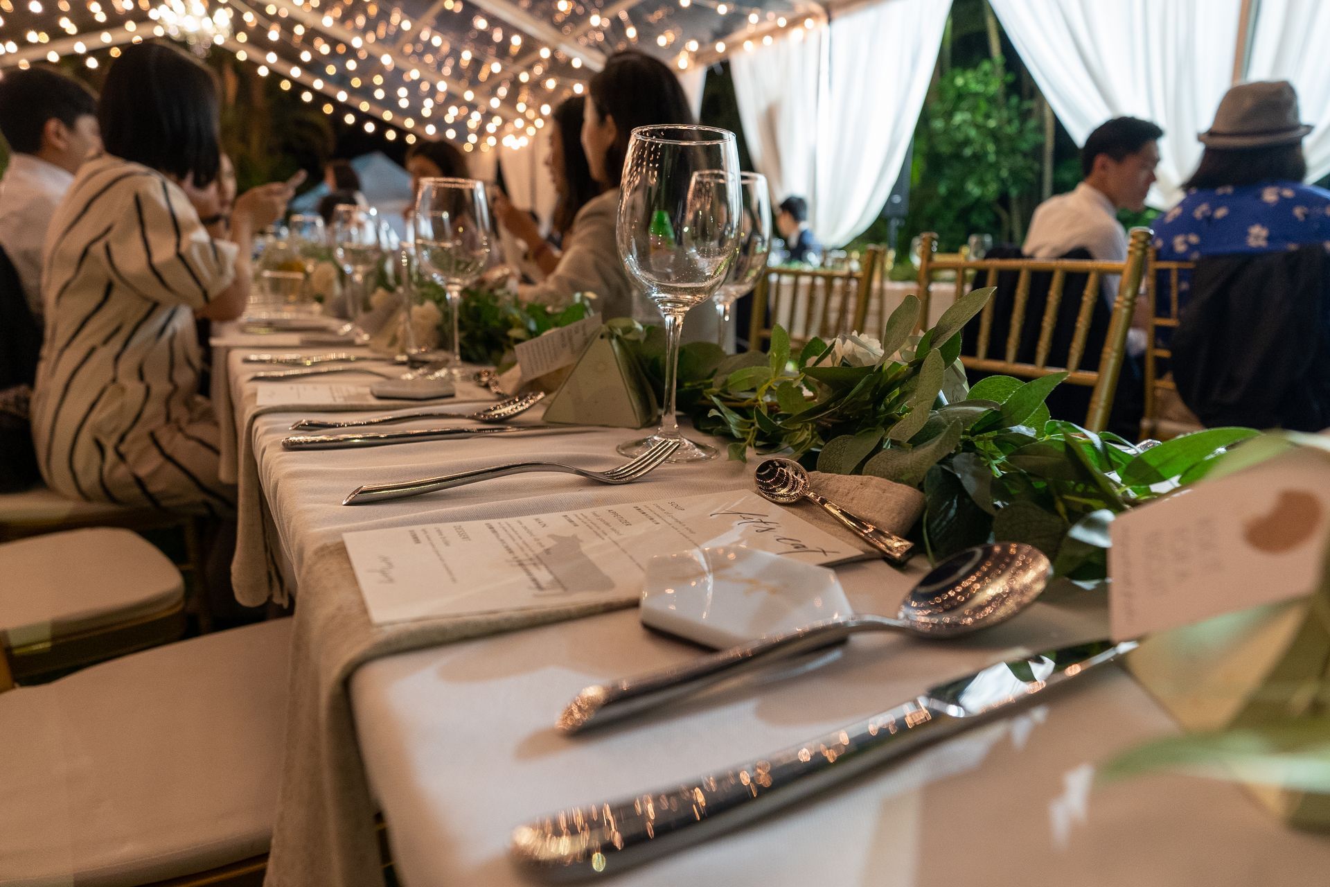 Long table set for an outdoor dinner, guests seated. Silverware, glasses, and greenery on the table under string lights.