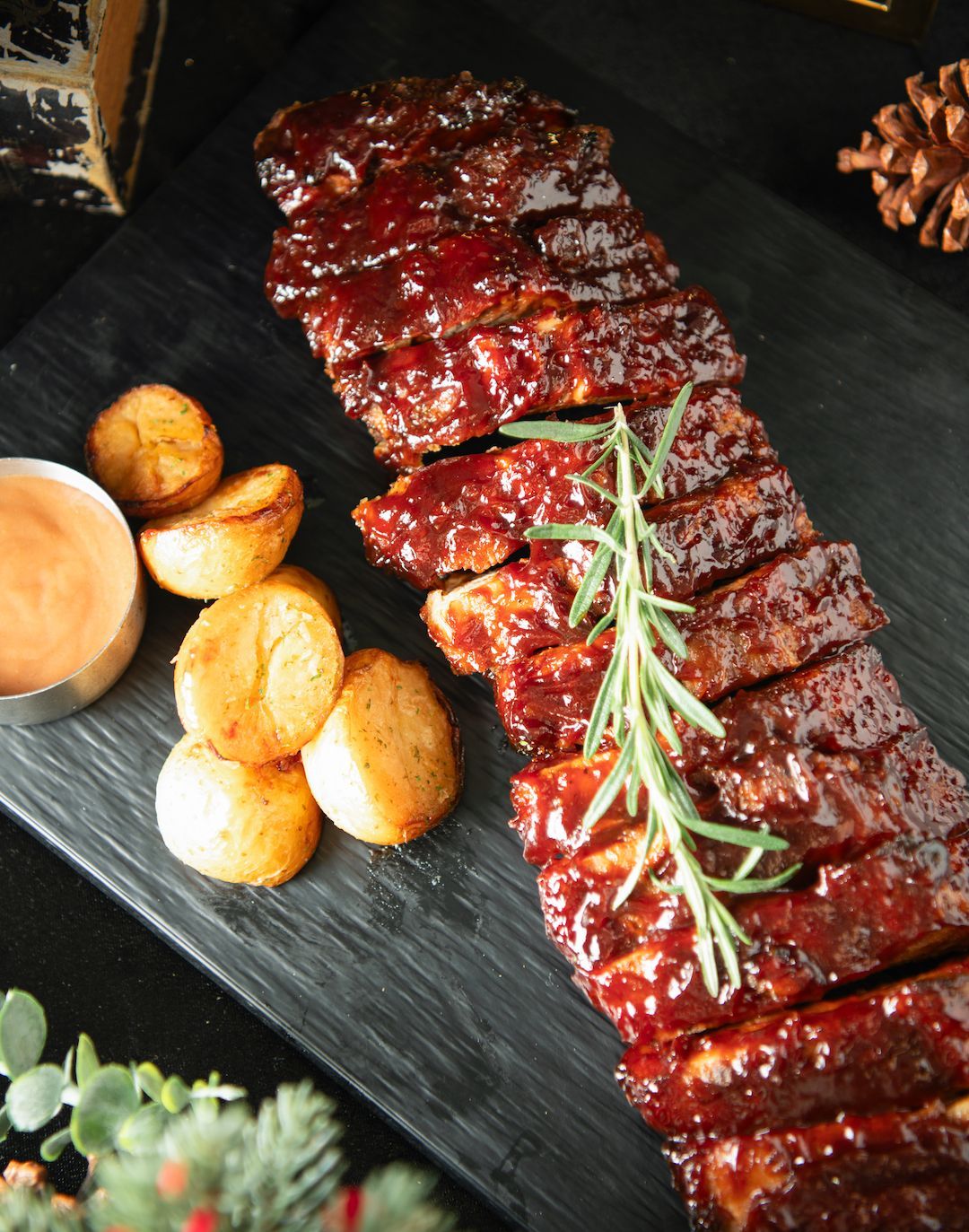 Rack of glazed ribs with roasted potatoes and dipping sauce, on a black plate.