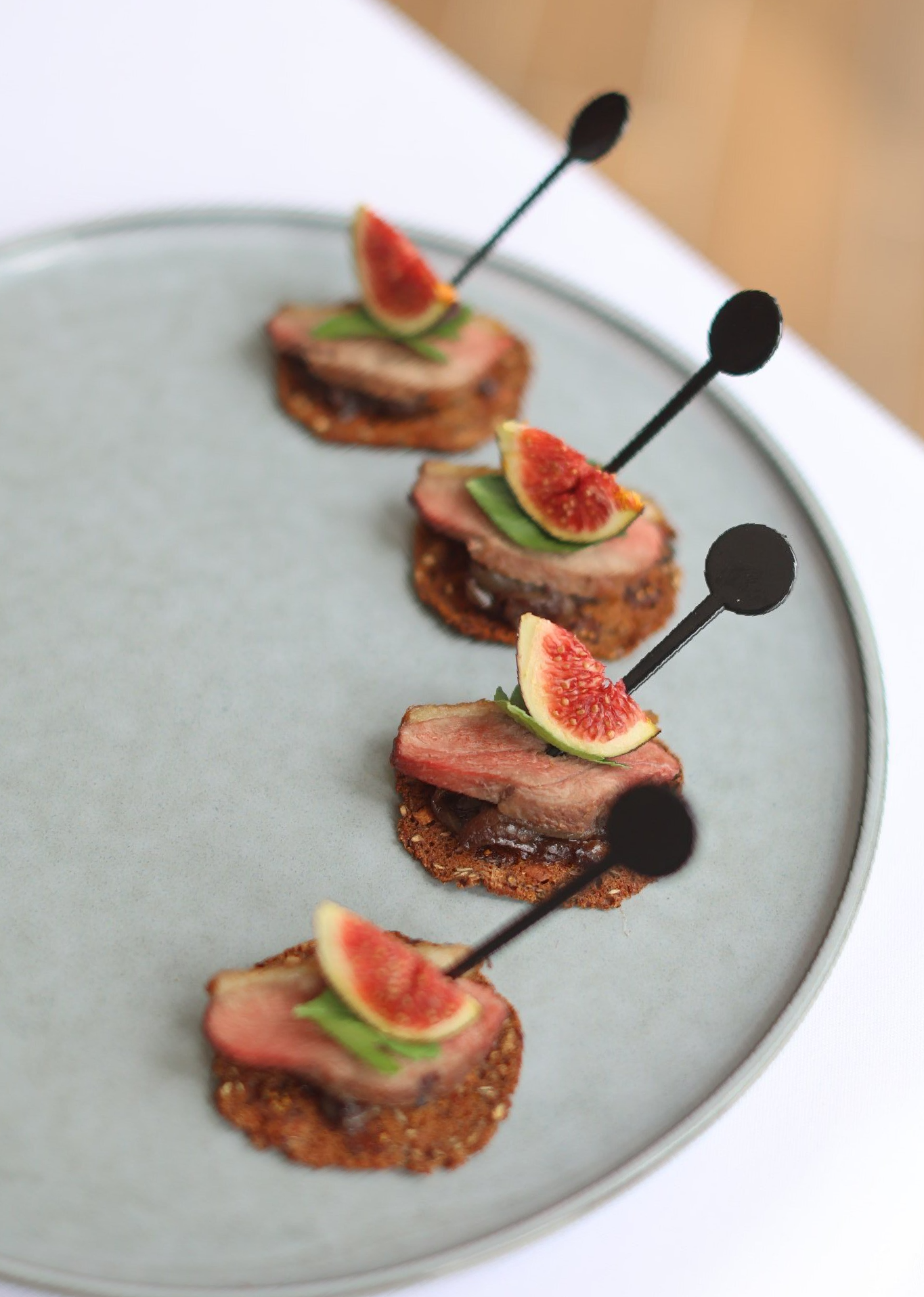 Four appetizers with meat, fig, and basil, served on a gray plate, each skewered with a black pick.