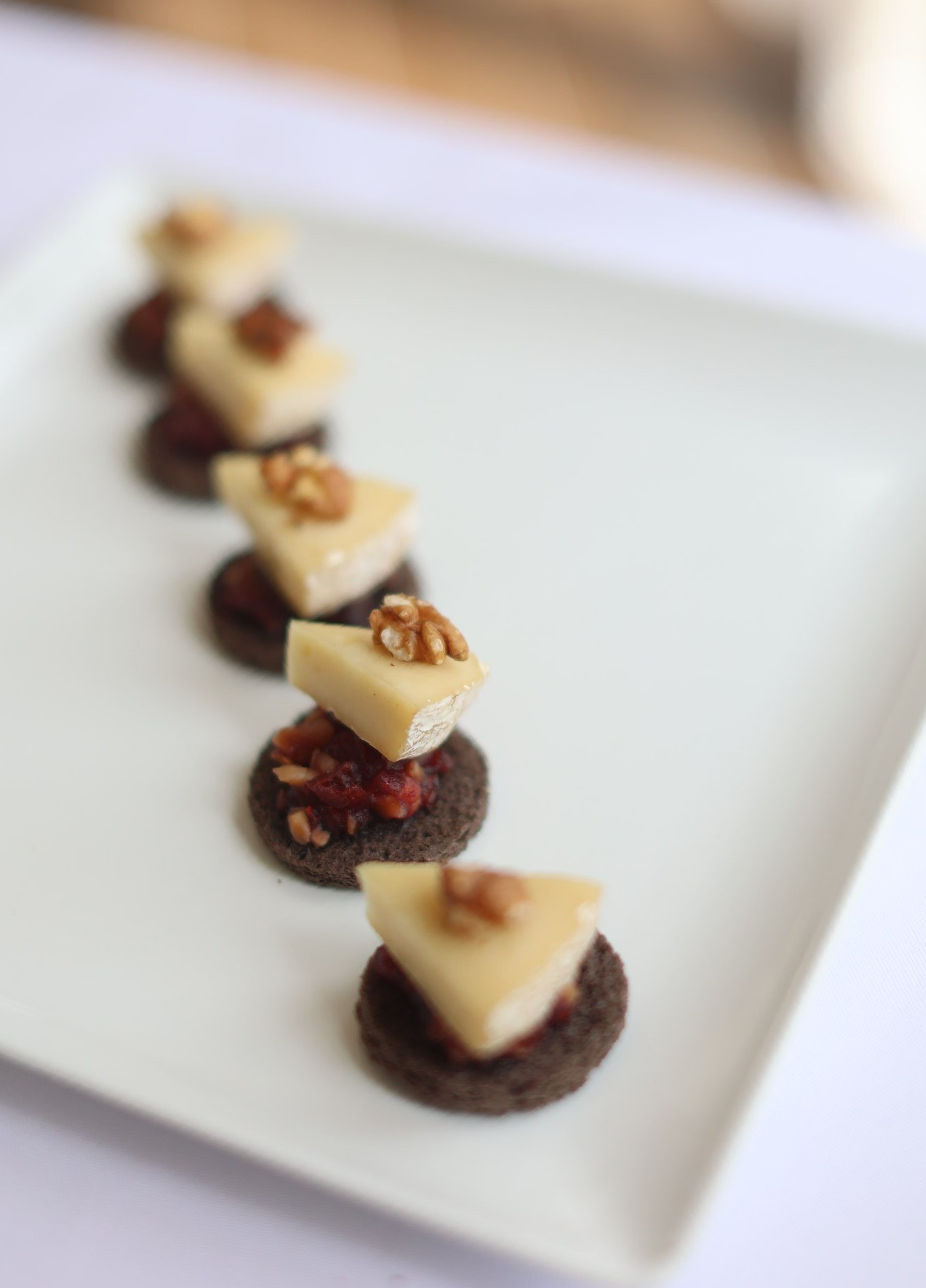 Appetizers: cheese triangles with walnuts and red topping on dark bread rounds, arranged on a white plate.