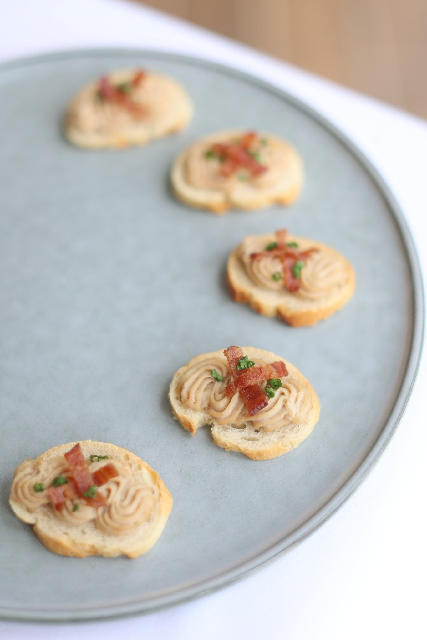 Appetizers on a round gray plate: toasted bread rounds topped with a light-colored spread and bacon bits.