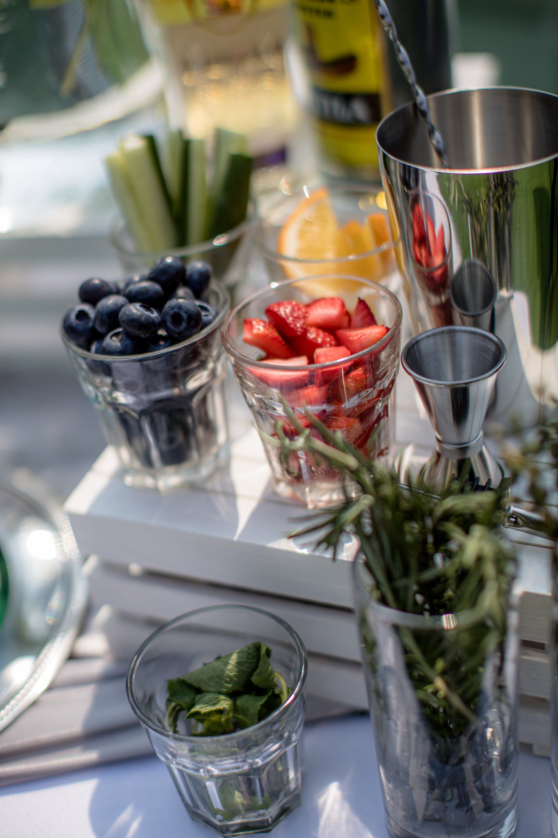 Close-up of a cocktail setup: blueberries, strawberries, orange slices, mint, cucumbers, rosemary, and bar tools.