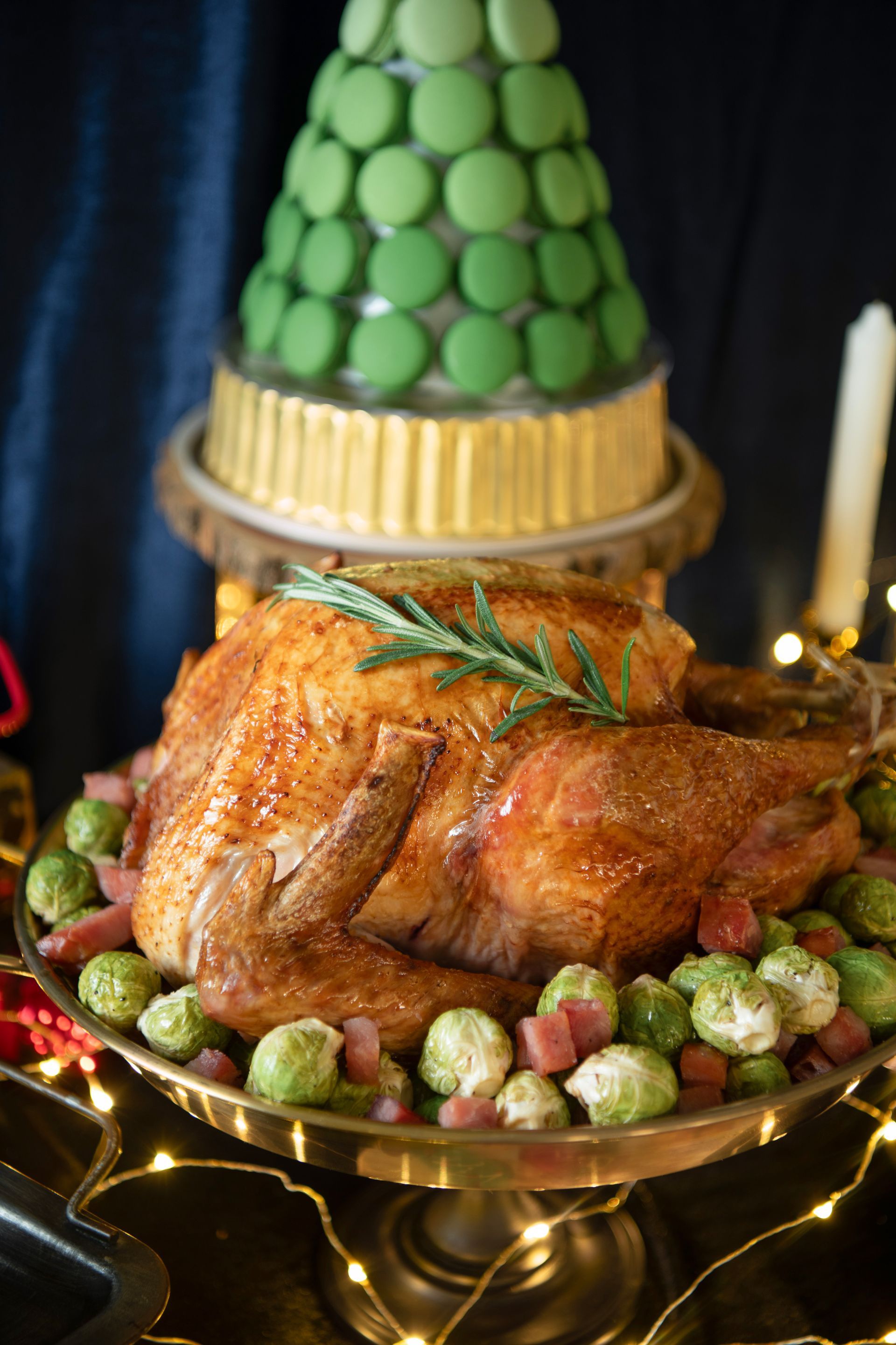 Roasted turkey on a platter with Brussels sprouts and a macaron tree cake in the background.