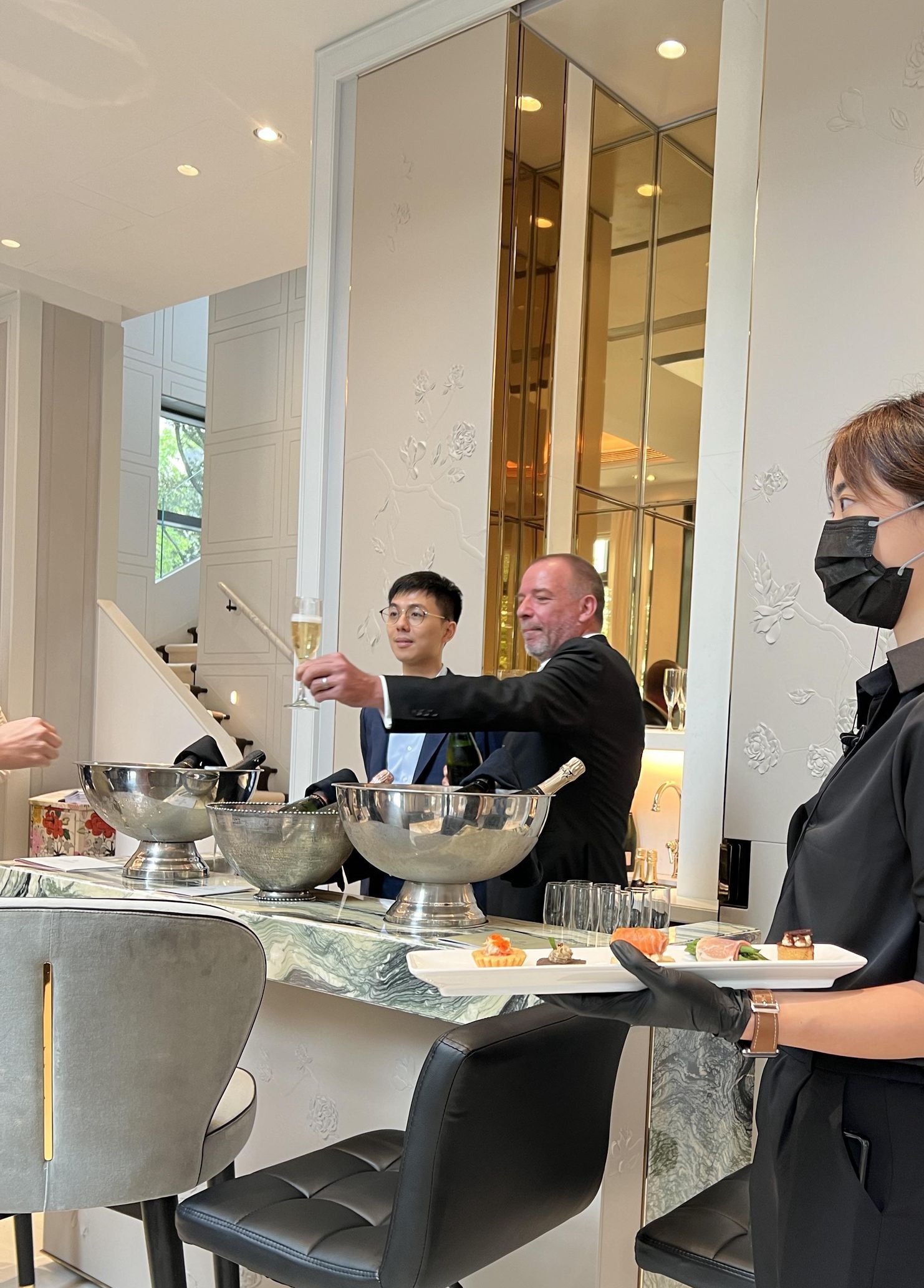 People toasting with champagne flutes near a marble bar. Servers with trays and a large mirror.