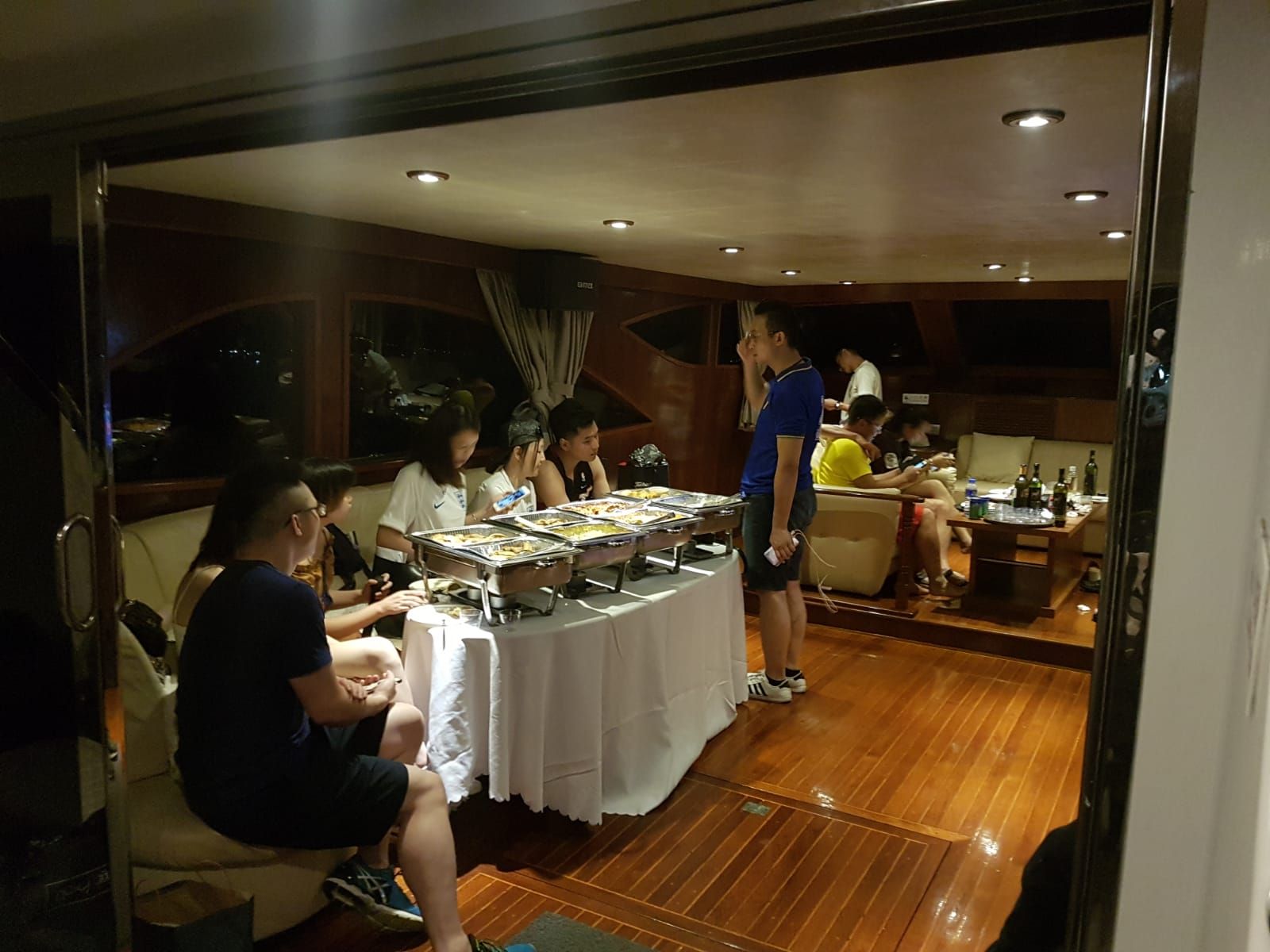 People at a buffet on a boat, inside a cabin. Wooden floor, food in silver containers, white tablecloth.