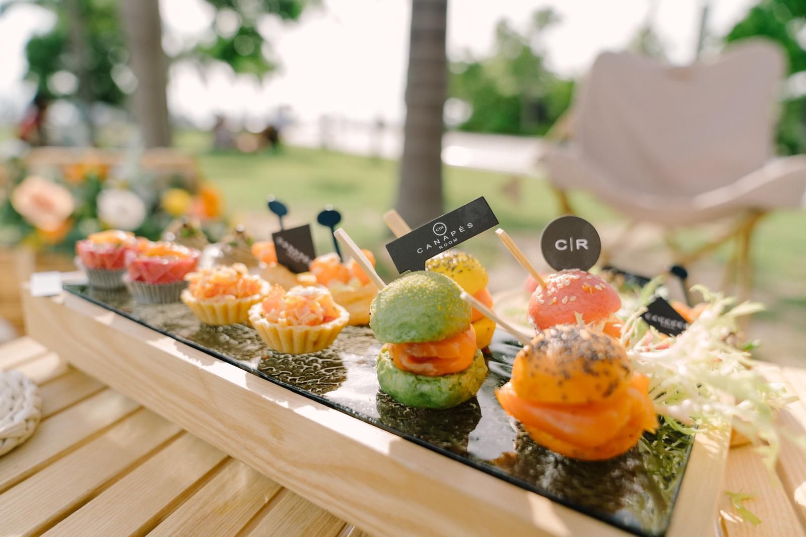 Appetizers on a wooden tray outdoors, including salmon bites and mini burgers.