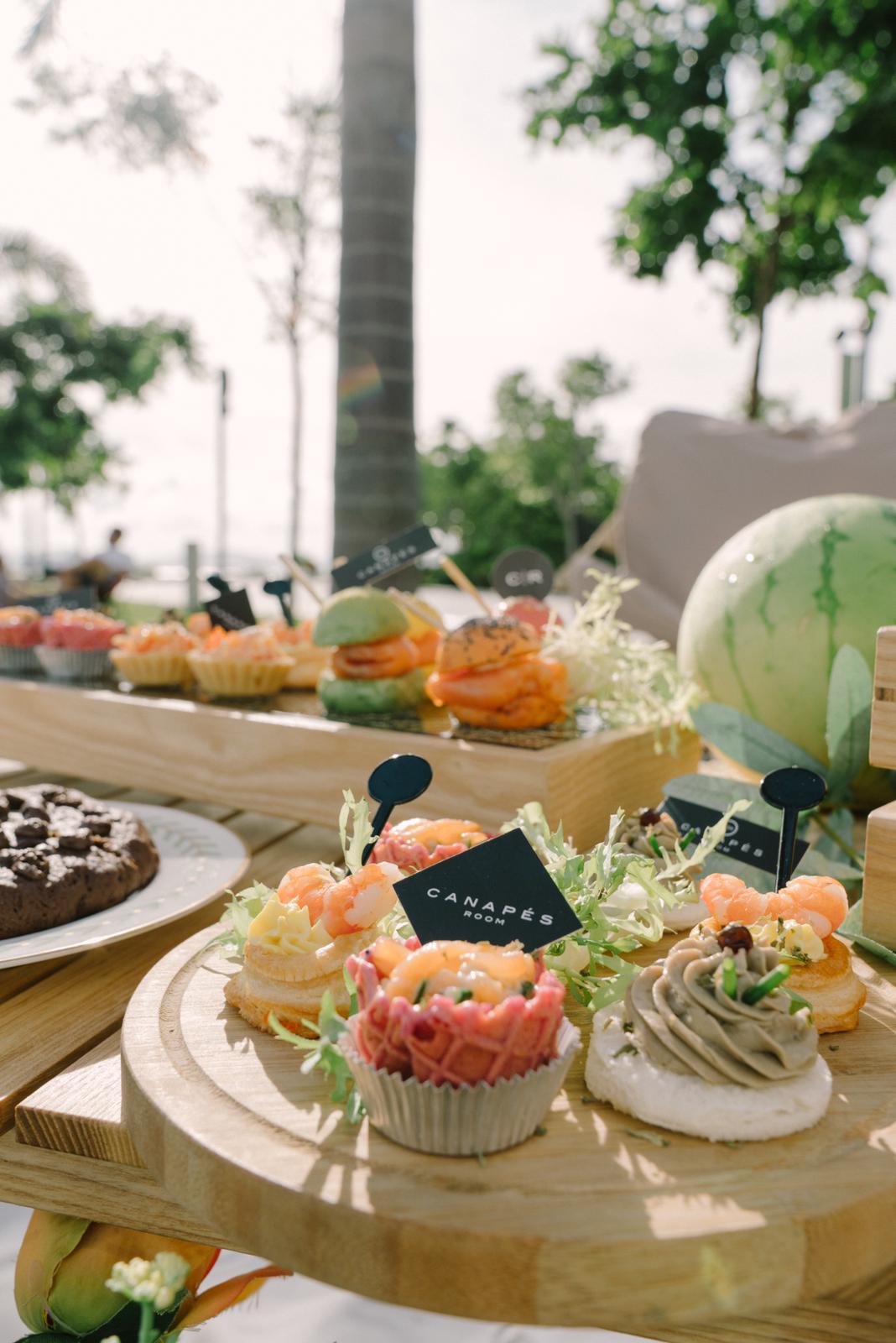 Assorted, colorful appetizers displayed on wooden platters, including mini sandwiches and pastries, outdoors.