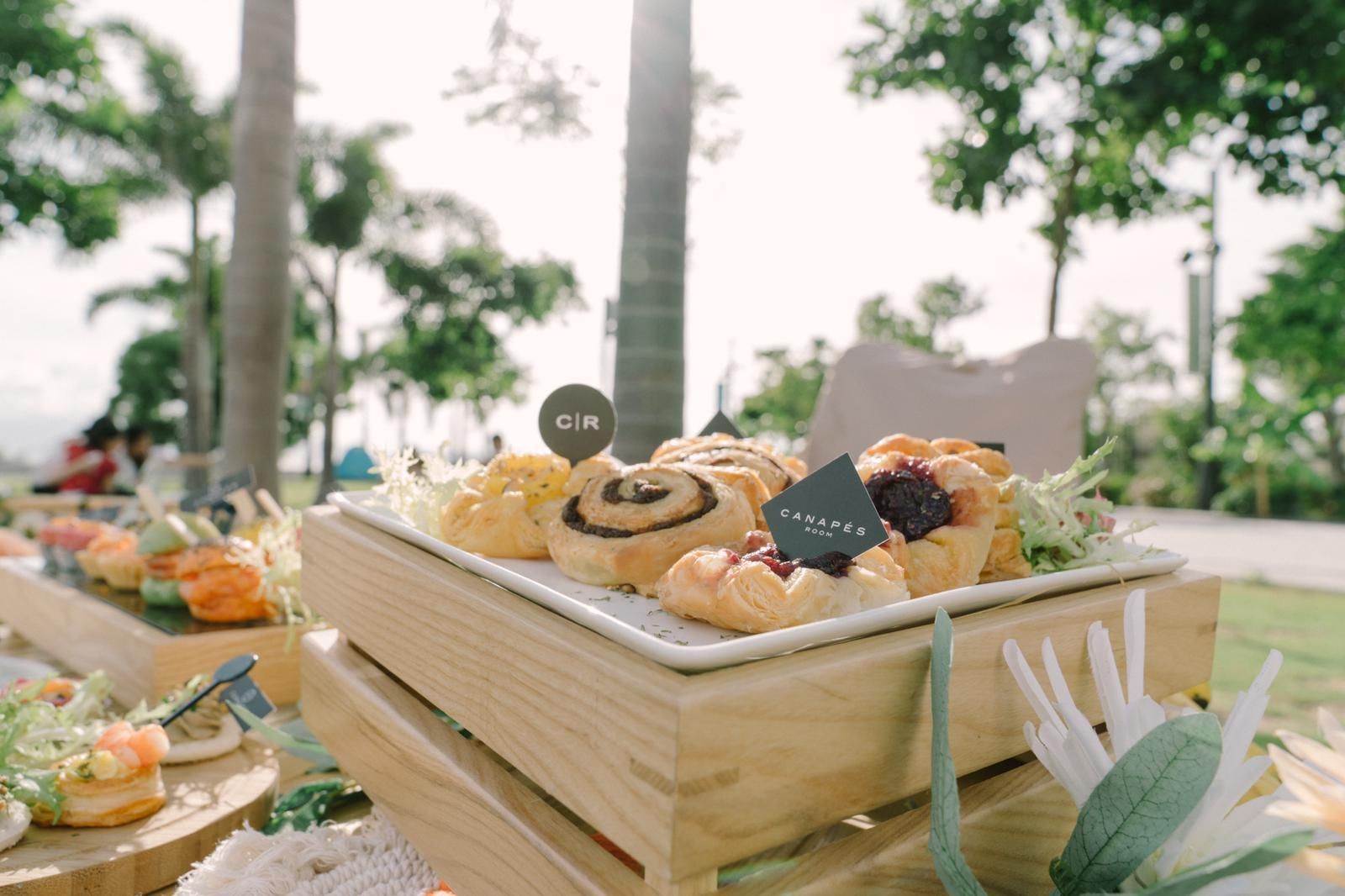 Pastries on a wooden stand at an outdoor event with greenery and a bright sky in the background.