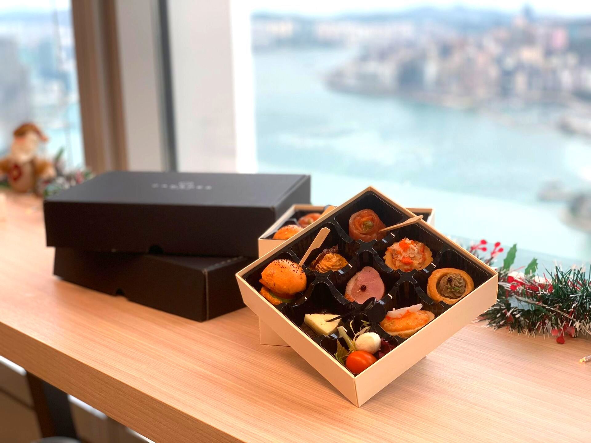 Open box of assorted appetizers on a wooden surface, with a city view in the background.