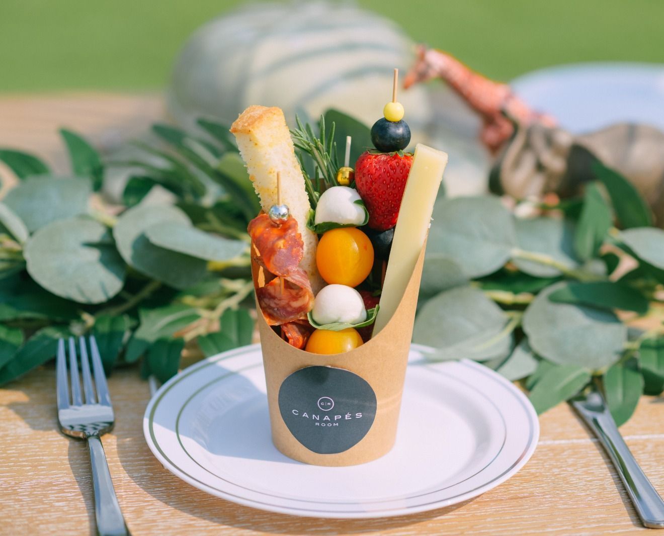 Skewered appetizers in a paper cup: vegetables, fruit, cheese, bread, and cured meats. Set outdoors with a fork and knife.