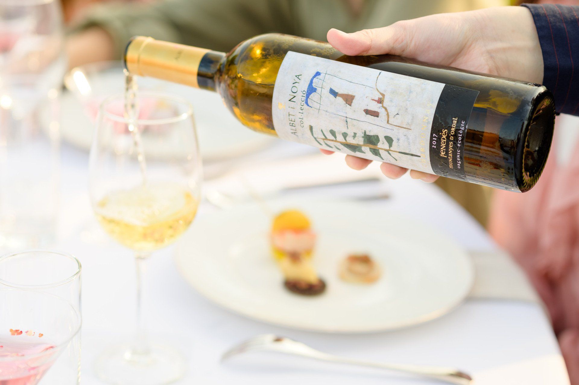 Person pouring white wine into a glass at a table setting, food in the background.
