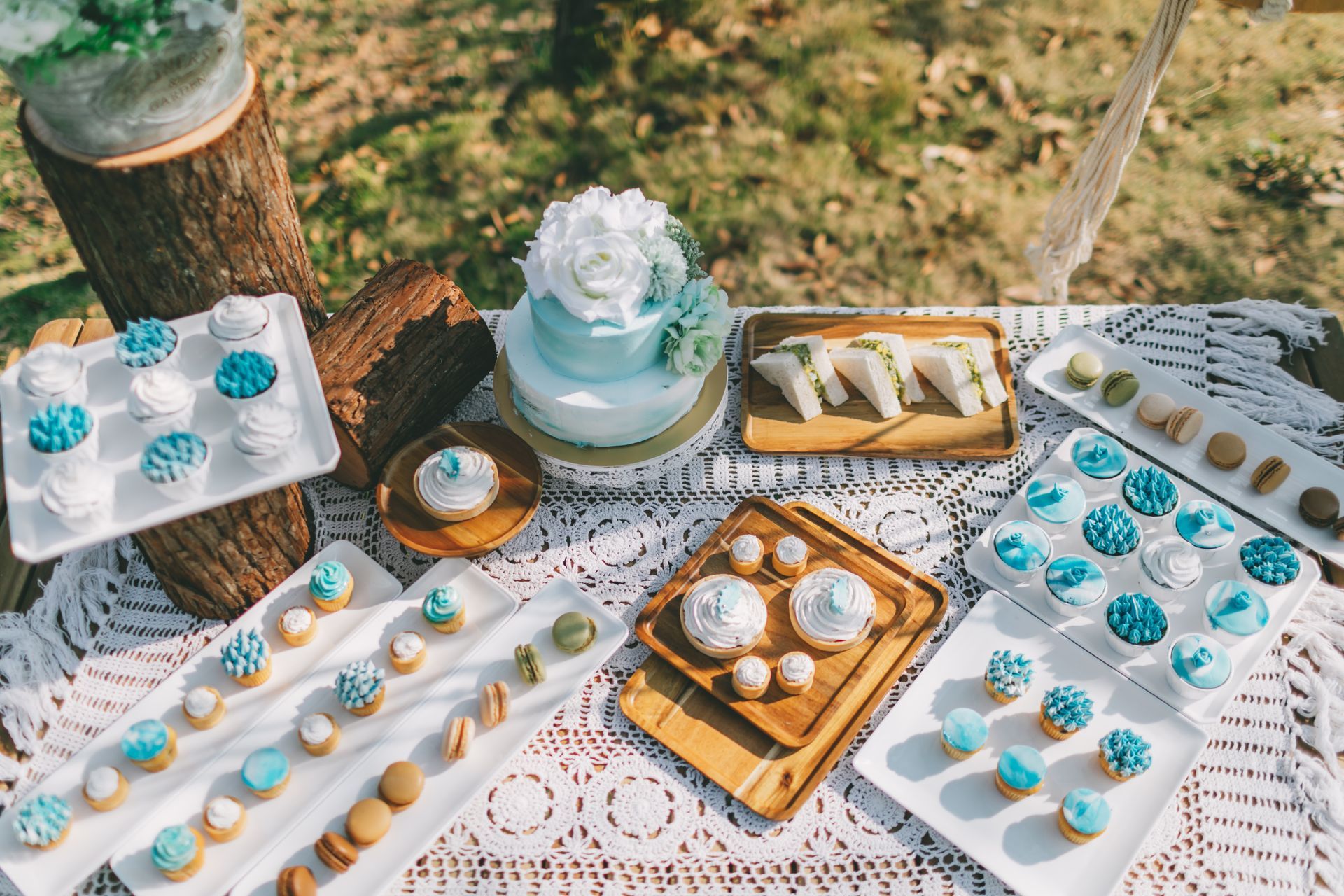 Dessert table with blue and white treats: cake, cupcakes, sandwiches, and cookies on a lace tablecloth in sunlight.
