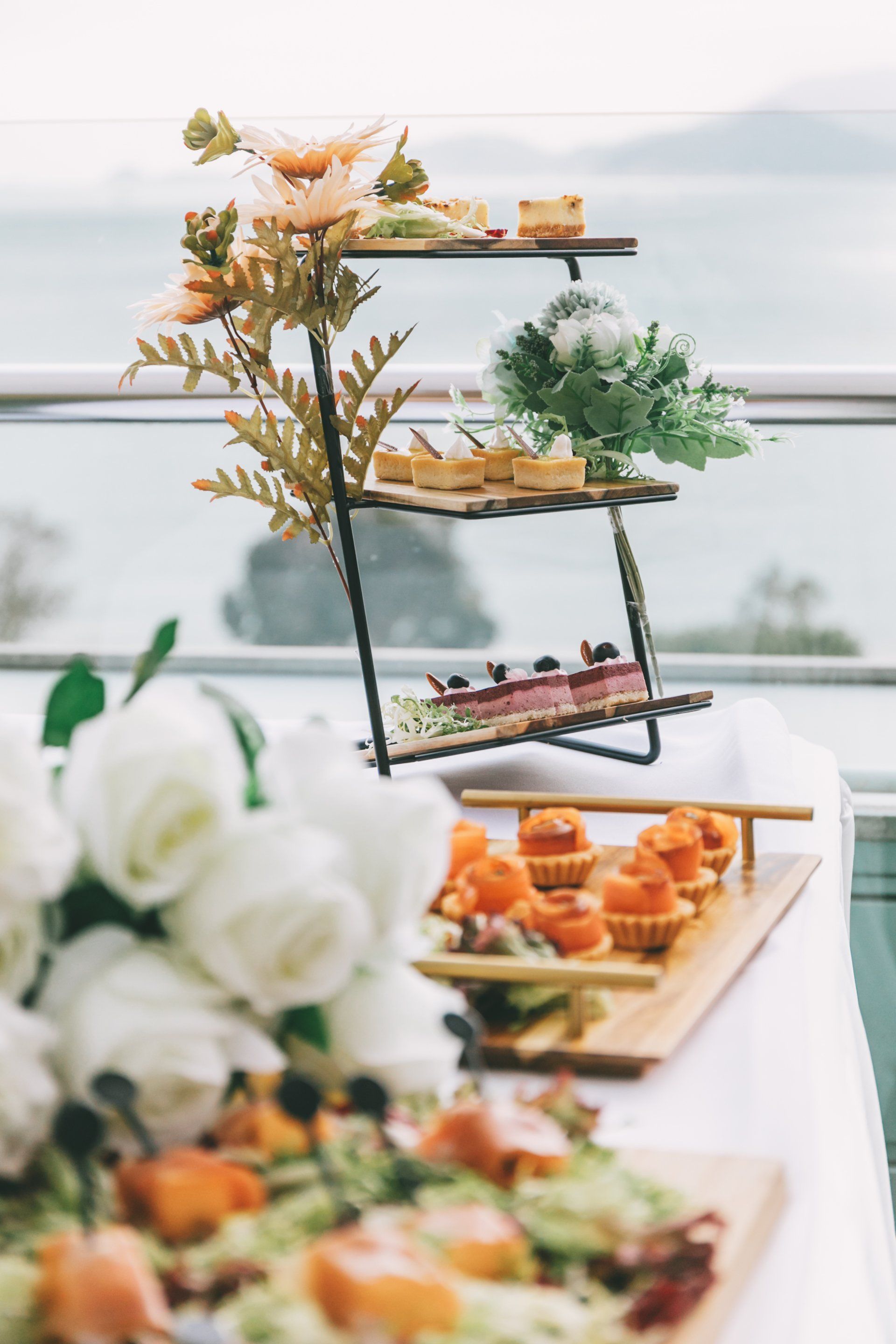 A three-tiered dessert stand with treats and flowers, set against a blurred seaside view.