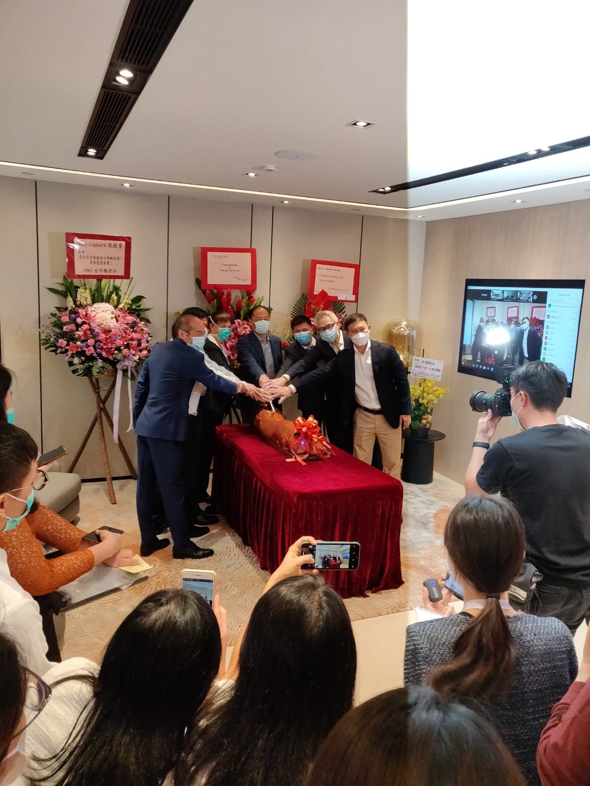 西裝革履的人們圍坐在一張鋪著紅色桌布的桌子旁,雙手伸向桌子。桌子上擺放著屏風和鮮花。