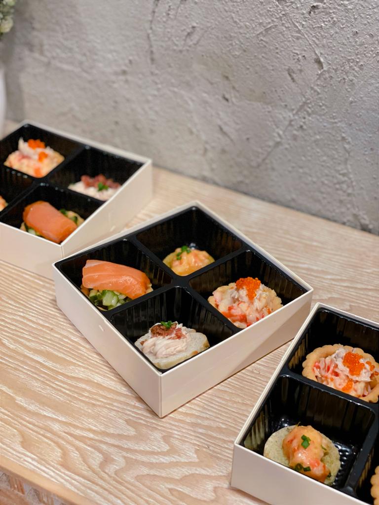 Sushi boxes with various toppings on a wooden surface against a textured gray wall.