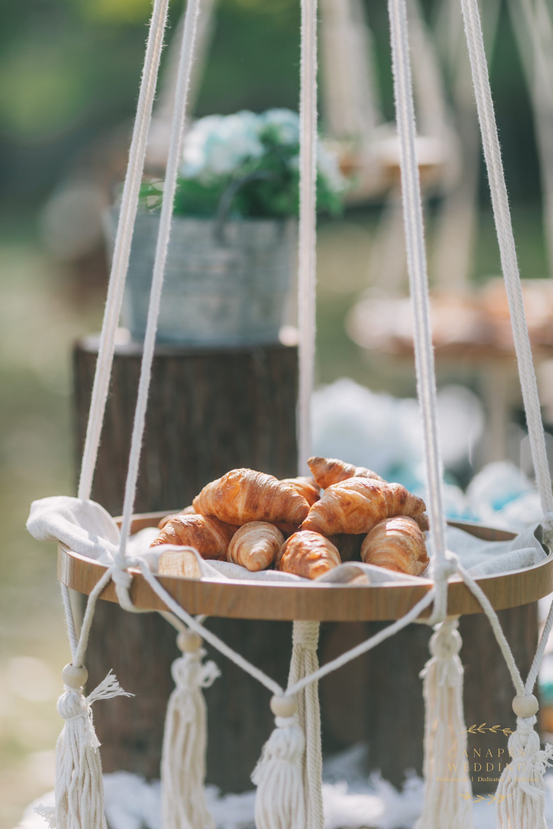 Croissants on a wooden tray suspended by macrame, set outdoors near a blurred background.