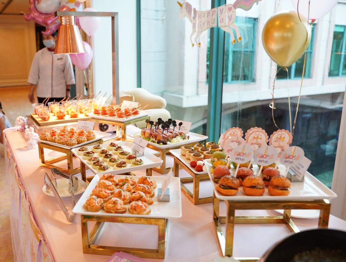 Buffet table with various appetizers, gold balloons, and a person in a chef coat.