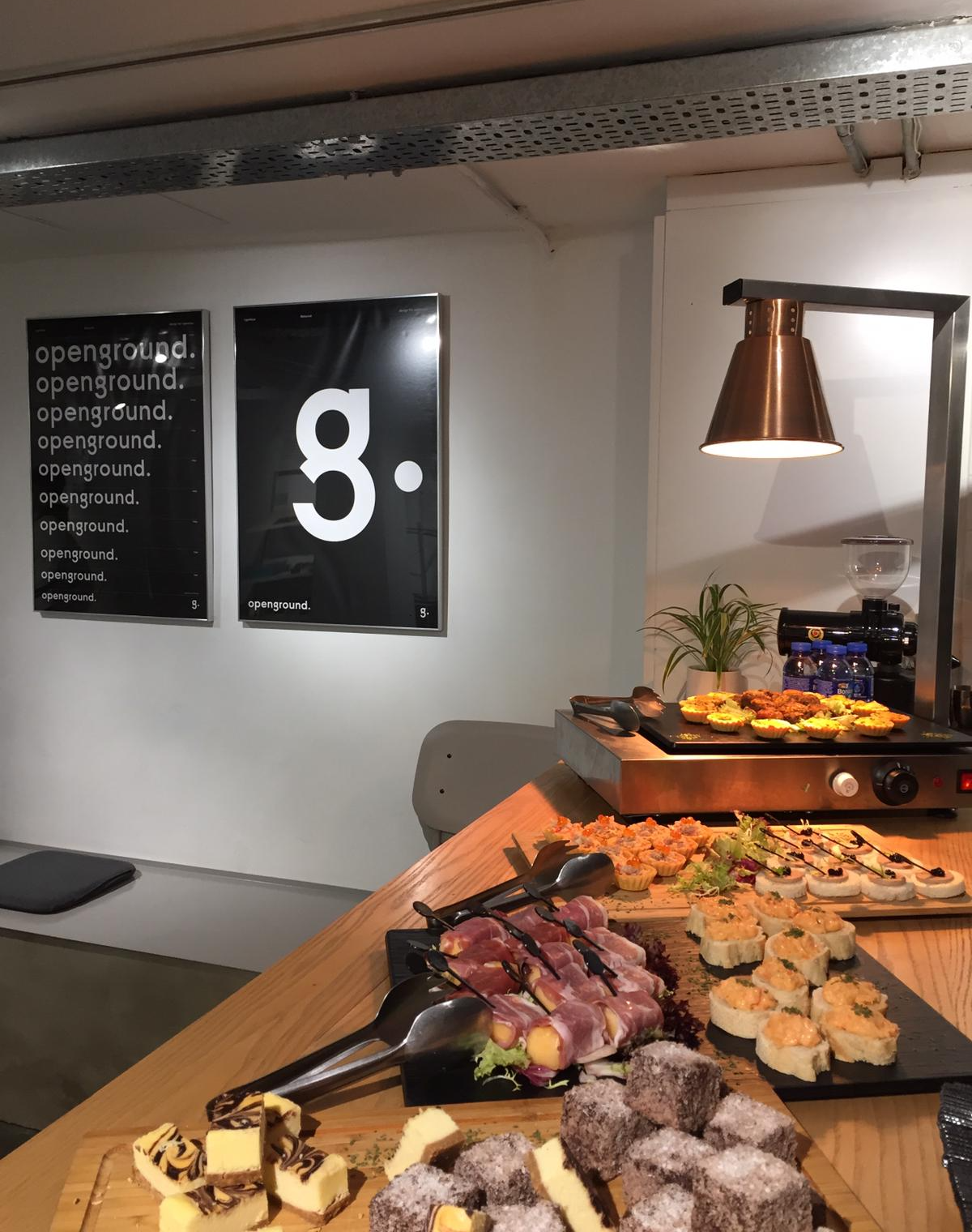 Catered food on a counter with a warmer, two black framed posters on a white wall.