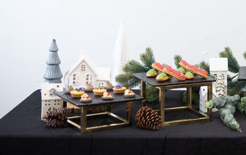 Dessert display: cakes on tiered stands, decorated with a pine cone, ceramic houses, and greenery on a black tablecloth.