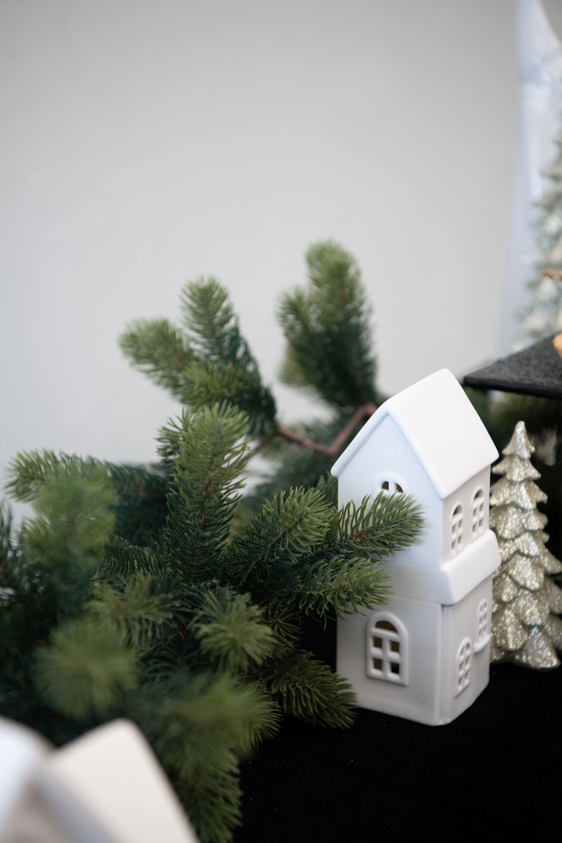 White ceramic house next to green fir branches and a silver Christmas tree.