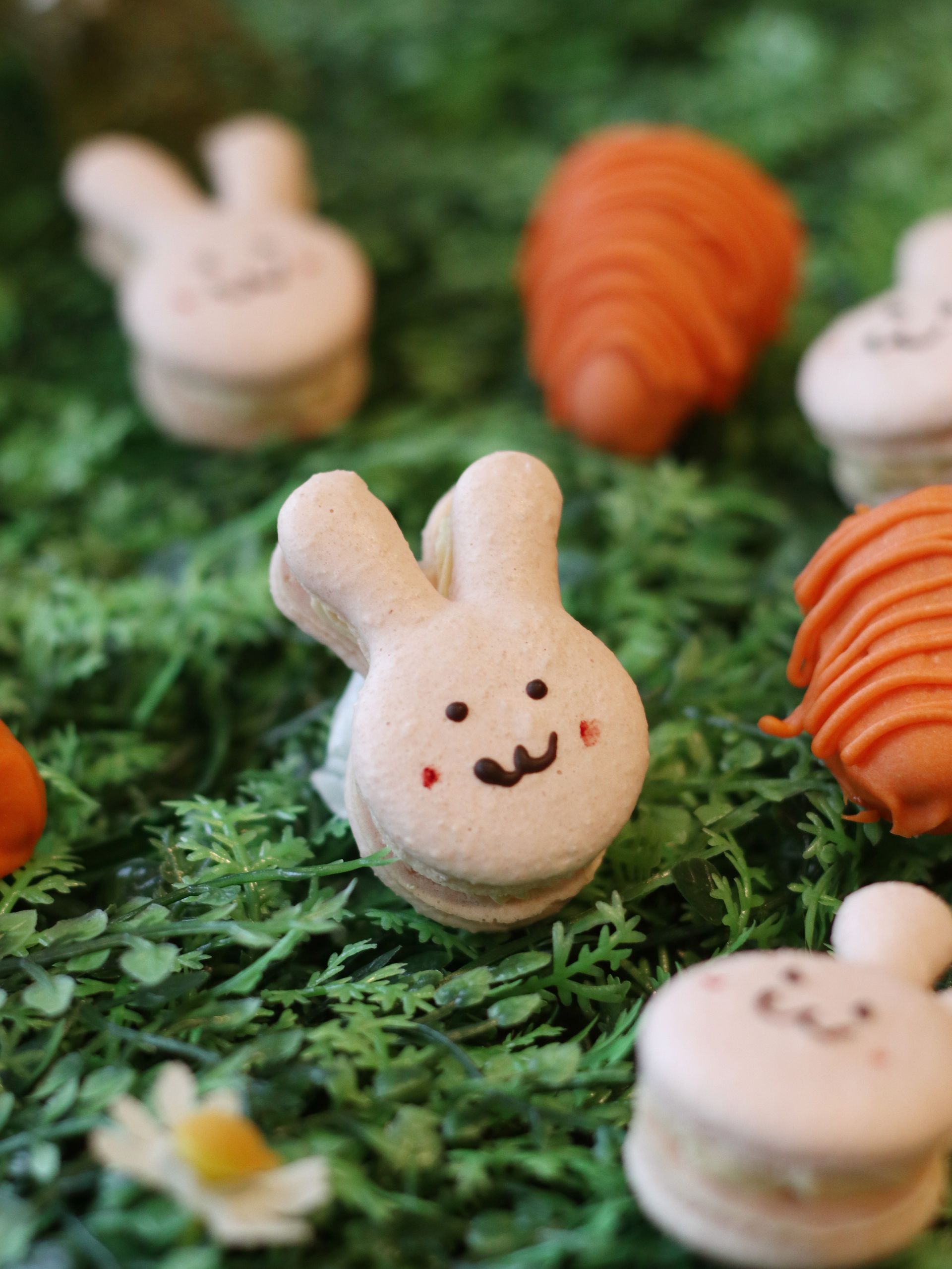 Bunny and carrot-shaped macarons on green artificial grass. White bunnies with smiling faces, orange carrots.