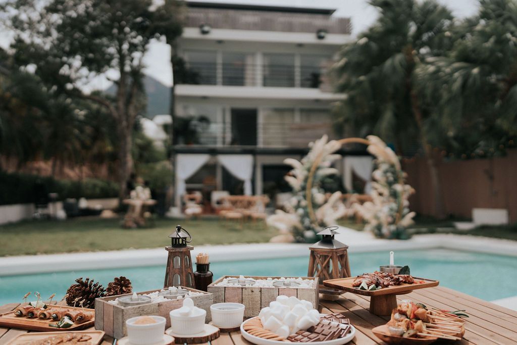 Outdoor wedding reception with pool, table of food and drinks, ceremony backdrop, and multi-story building.