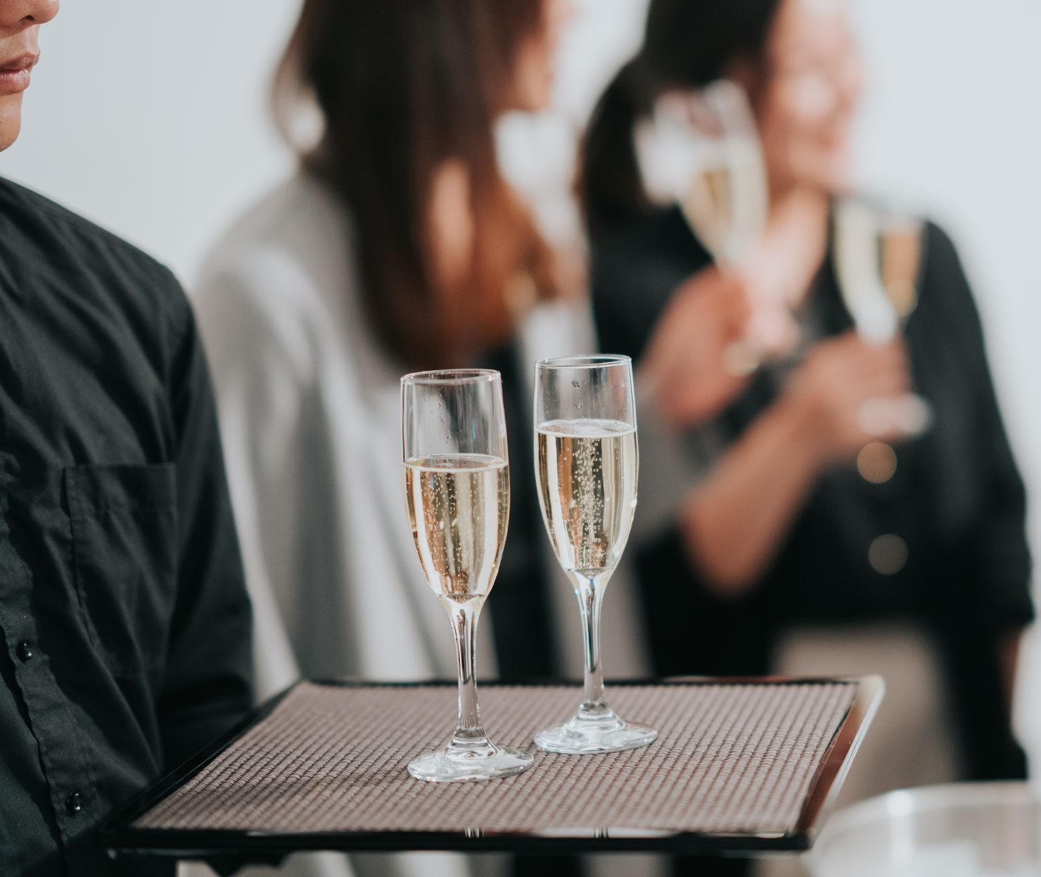 Waiter holding a tray with two champagne flutes, other guests blurred in the background.