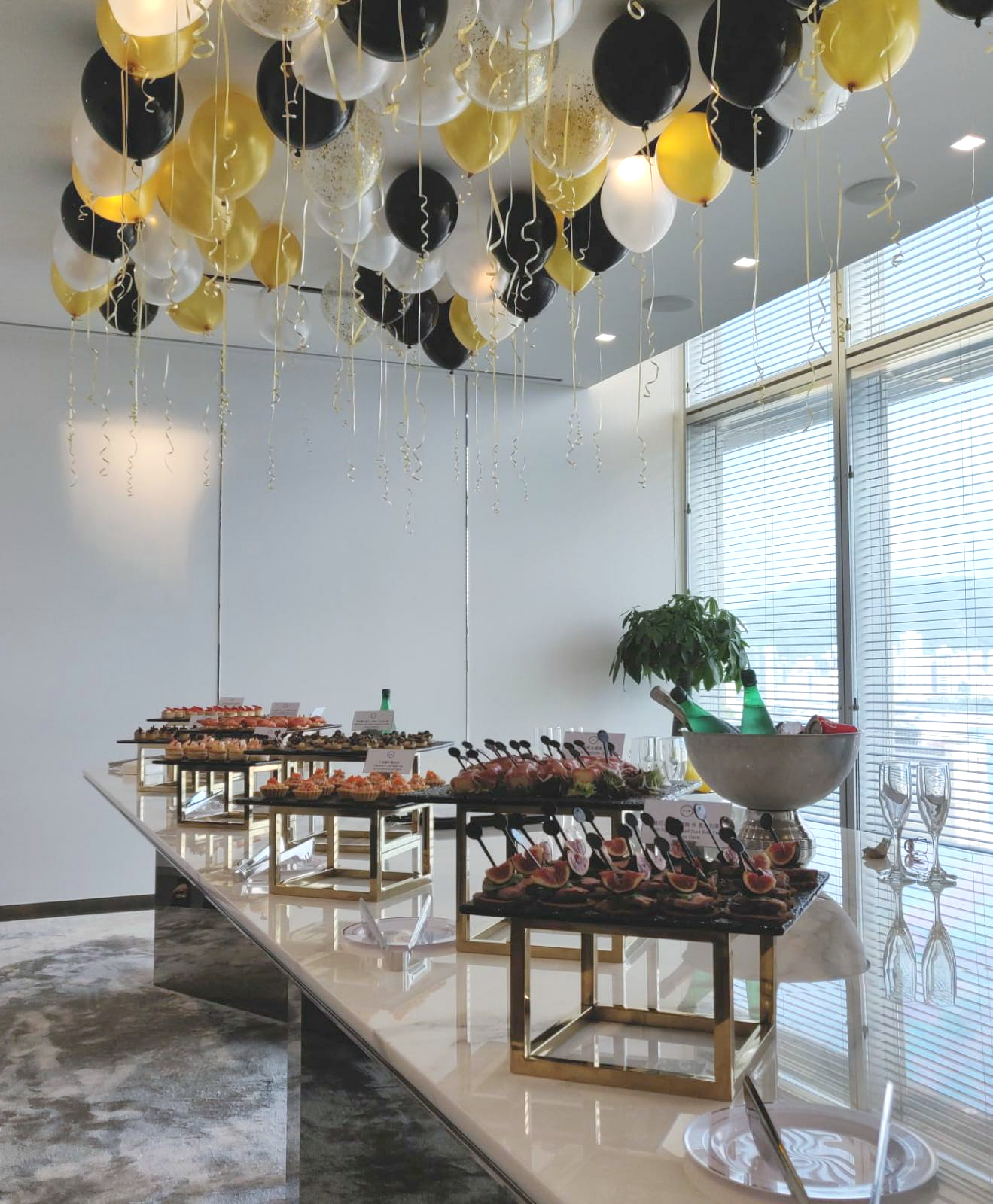 Buffet table with hors d'oeuvres under gold, black, and white balloons. Indoor event setting.