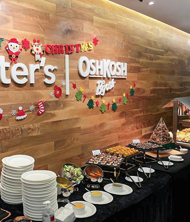 Christmas buffet with Carter's Oshkosh logo on wood-paneled wall, plates, and various food trays.
