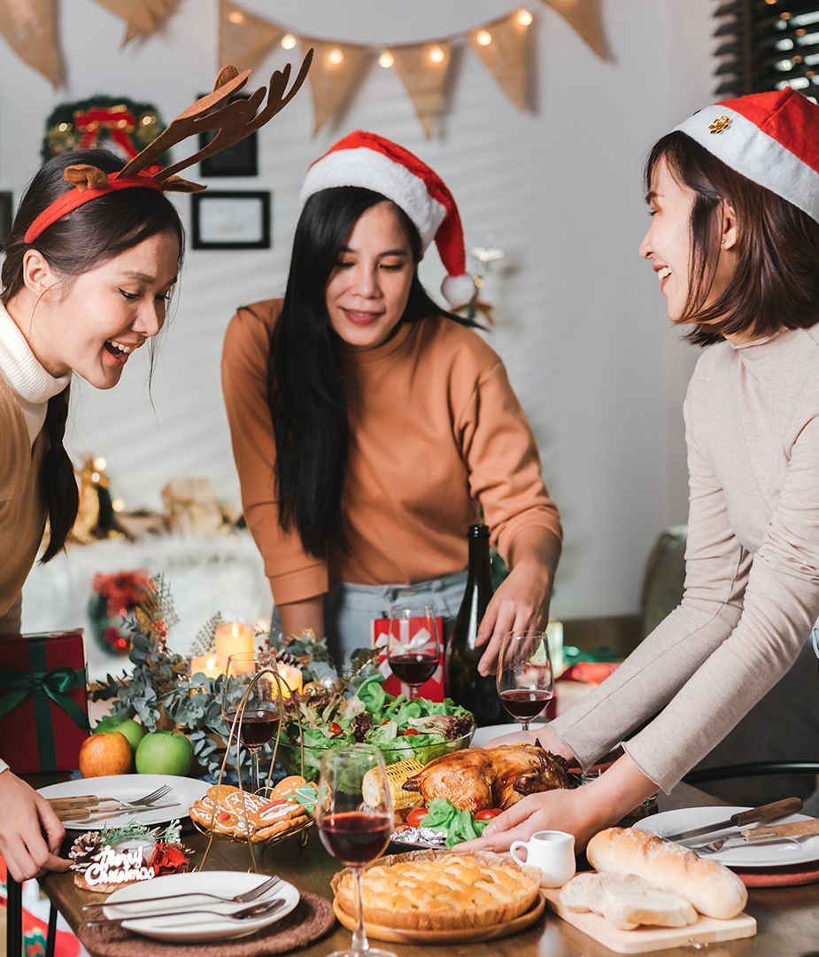 Three women preparing a Christmas dinner, smiling. Table set with food, wine, and decorations.