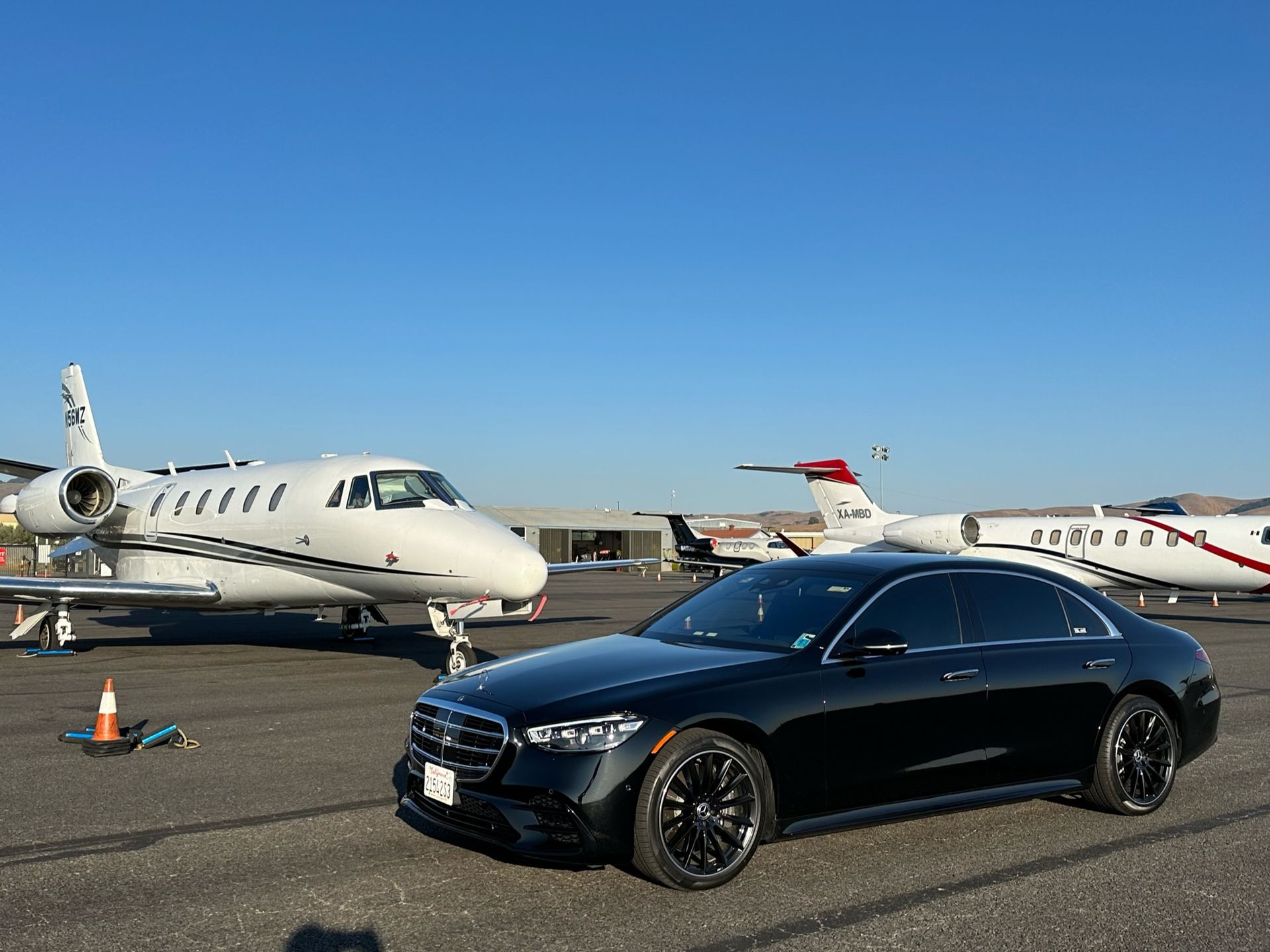 Black car parked on tarmac with private jets under a blue sky.