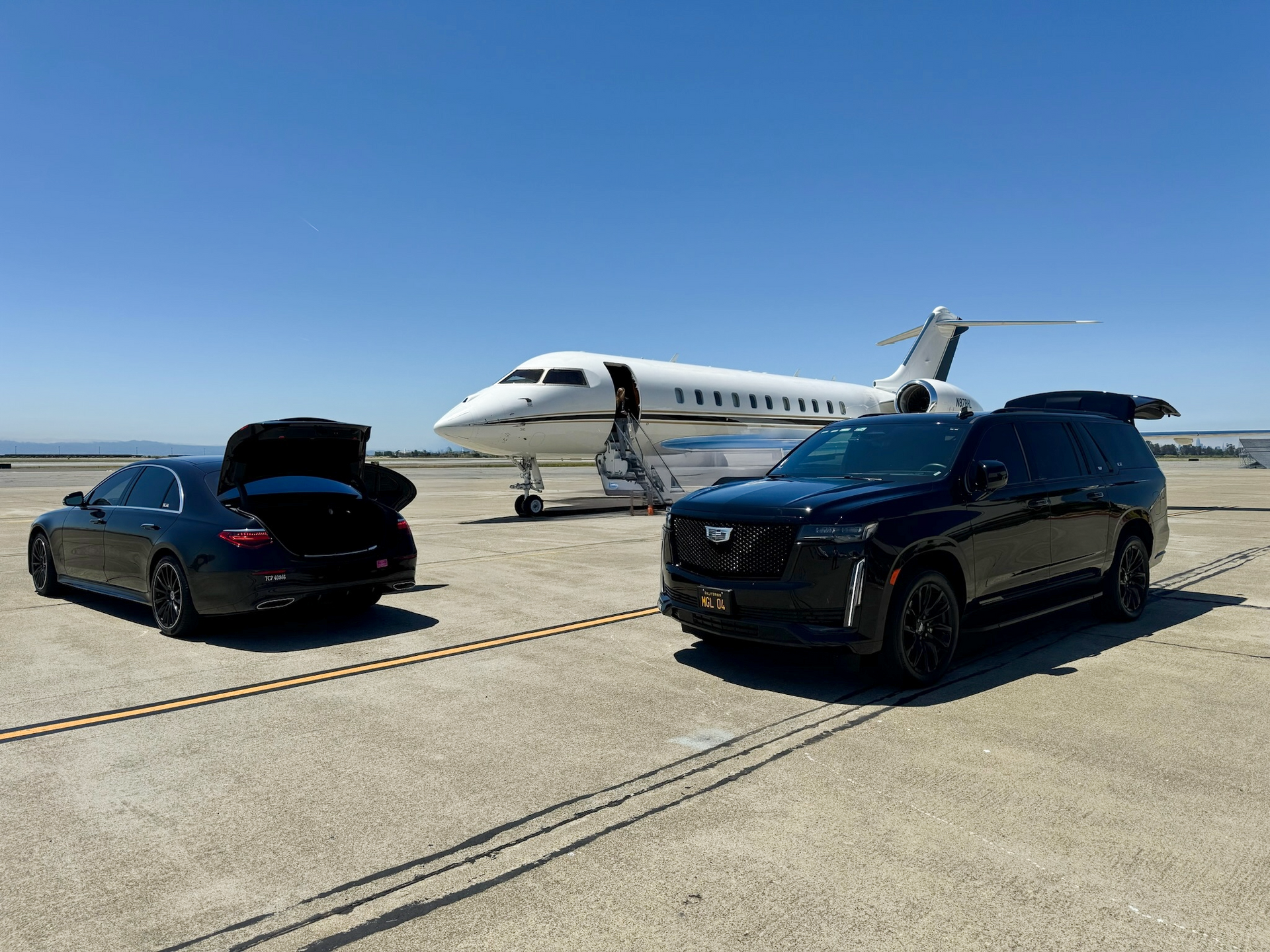 Private jet on tarmac with  black SUV and sedan, one with its trunk open under a clear blue sky