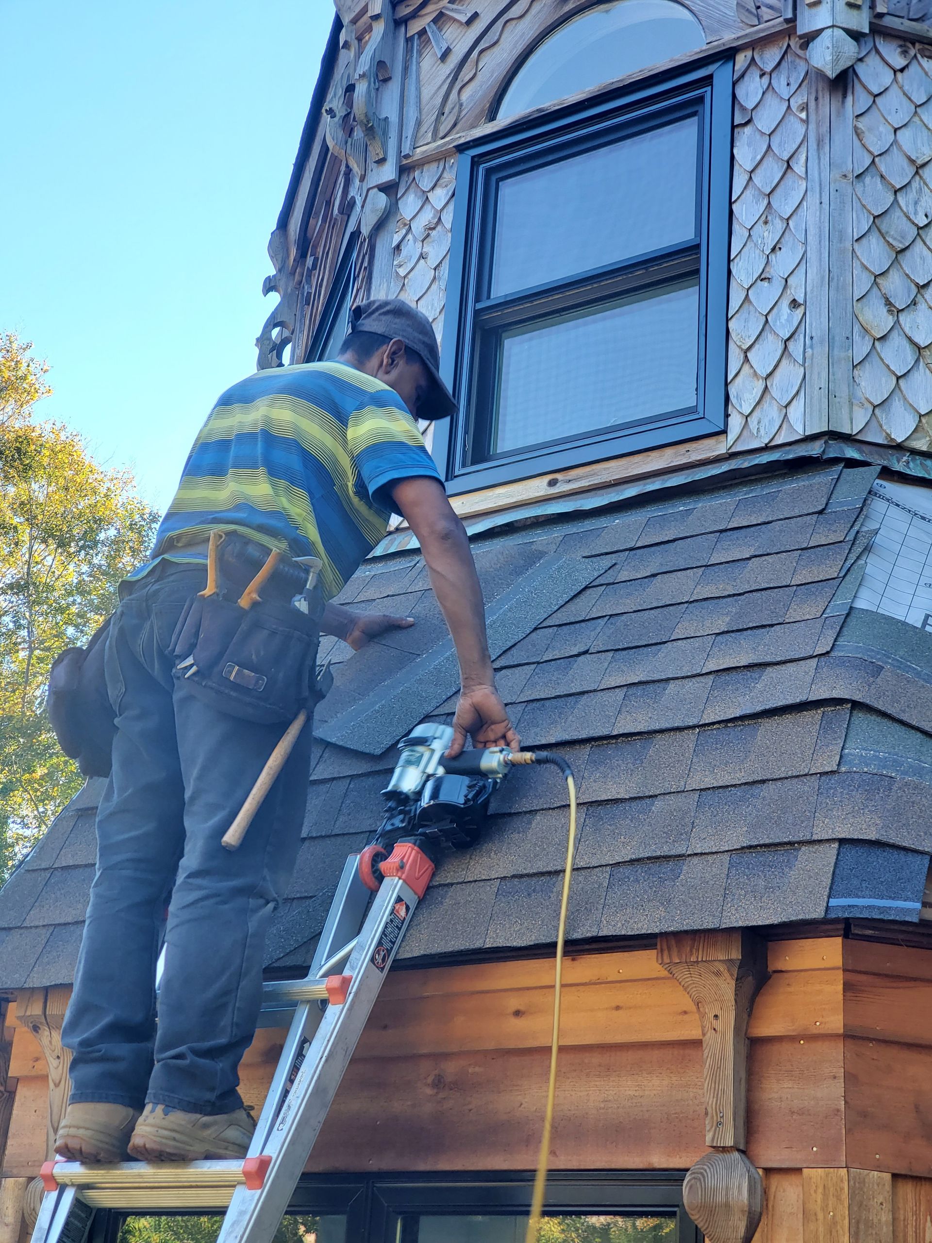 A construction worker, standing on a ladder, works on a shingle roof next to a window. 