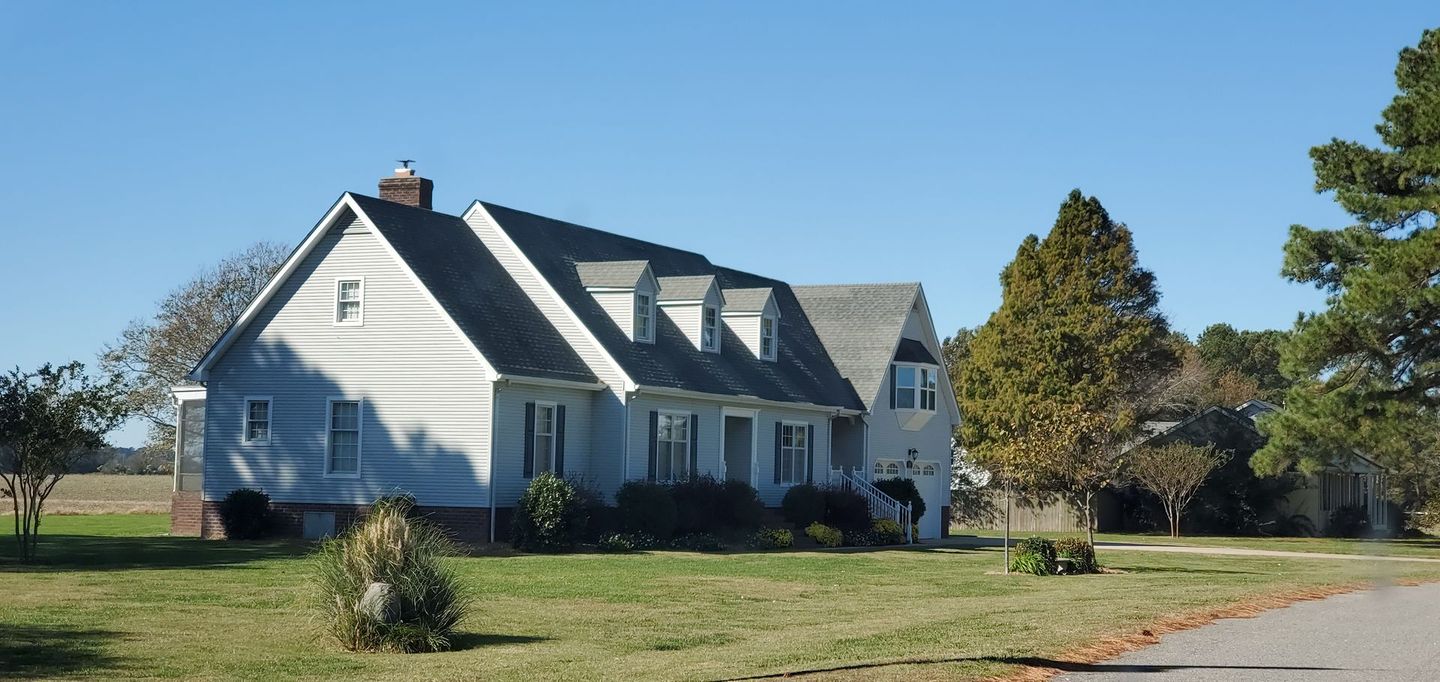 A two-story gray house with a dark roof sits on a grassy lawn under a clear blue sky.  Trees border the property.