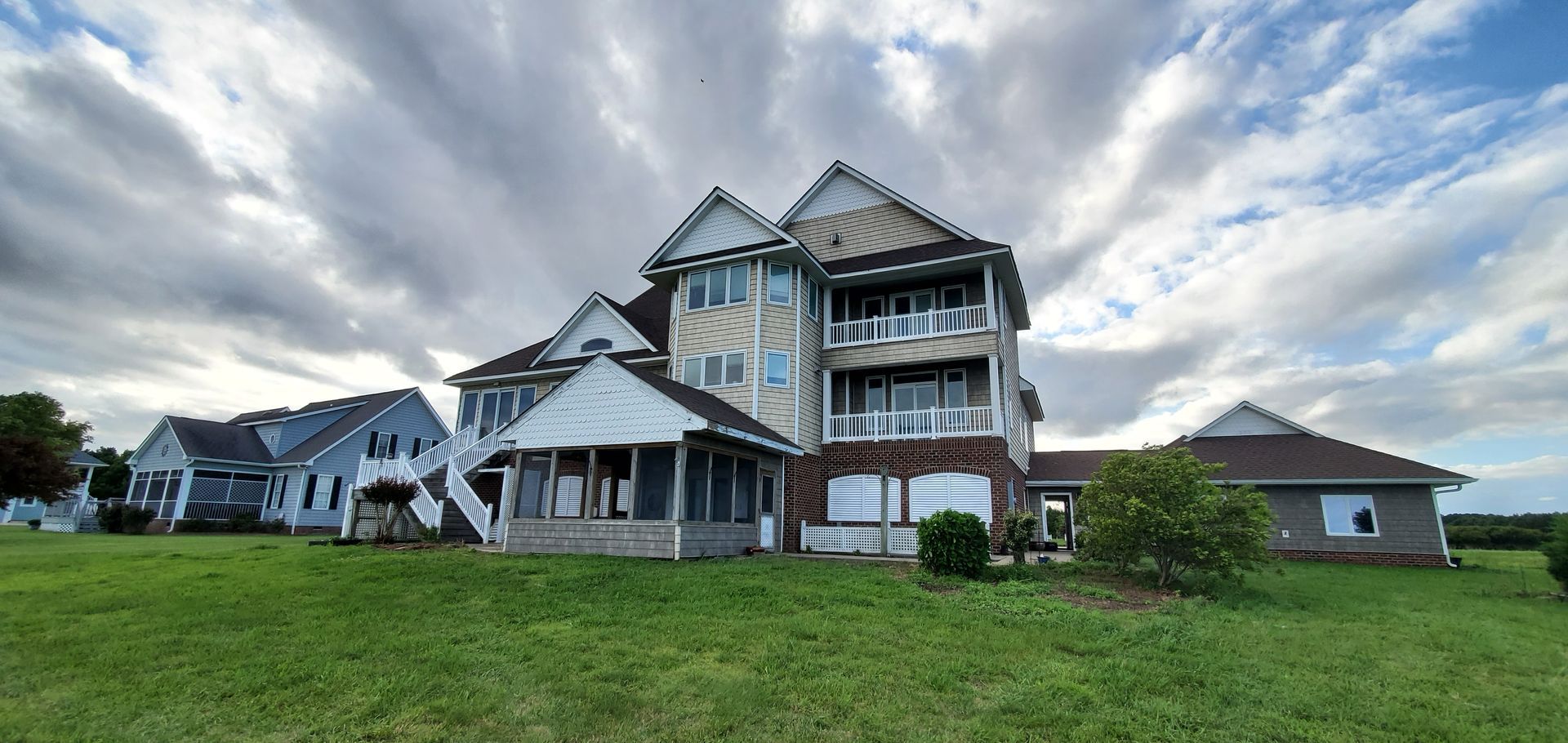 A multi-story house with balconies stands on a grassy lawn under a cloudy sky. Other houses are visible in the background.