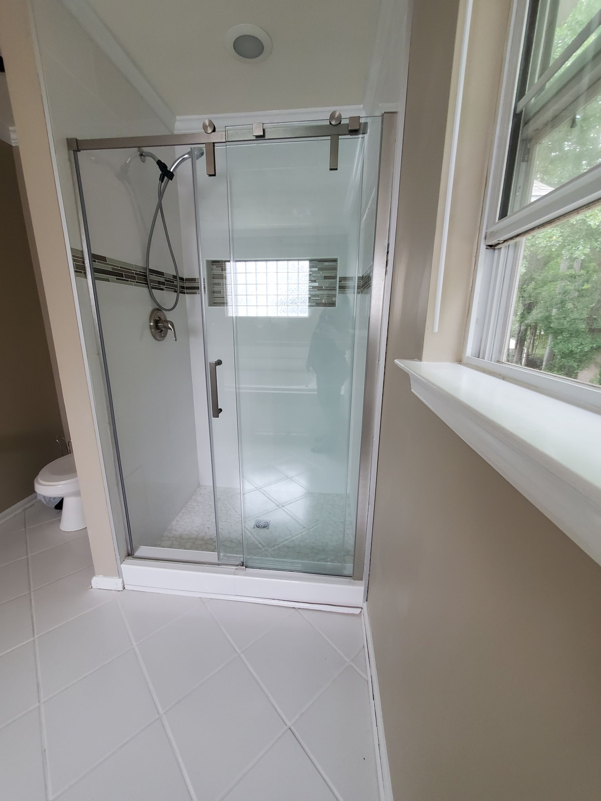 A modern bathroom with a glass shower enclosure, white tile floor, and a window to the right.