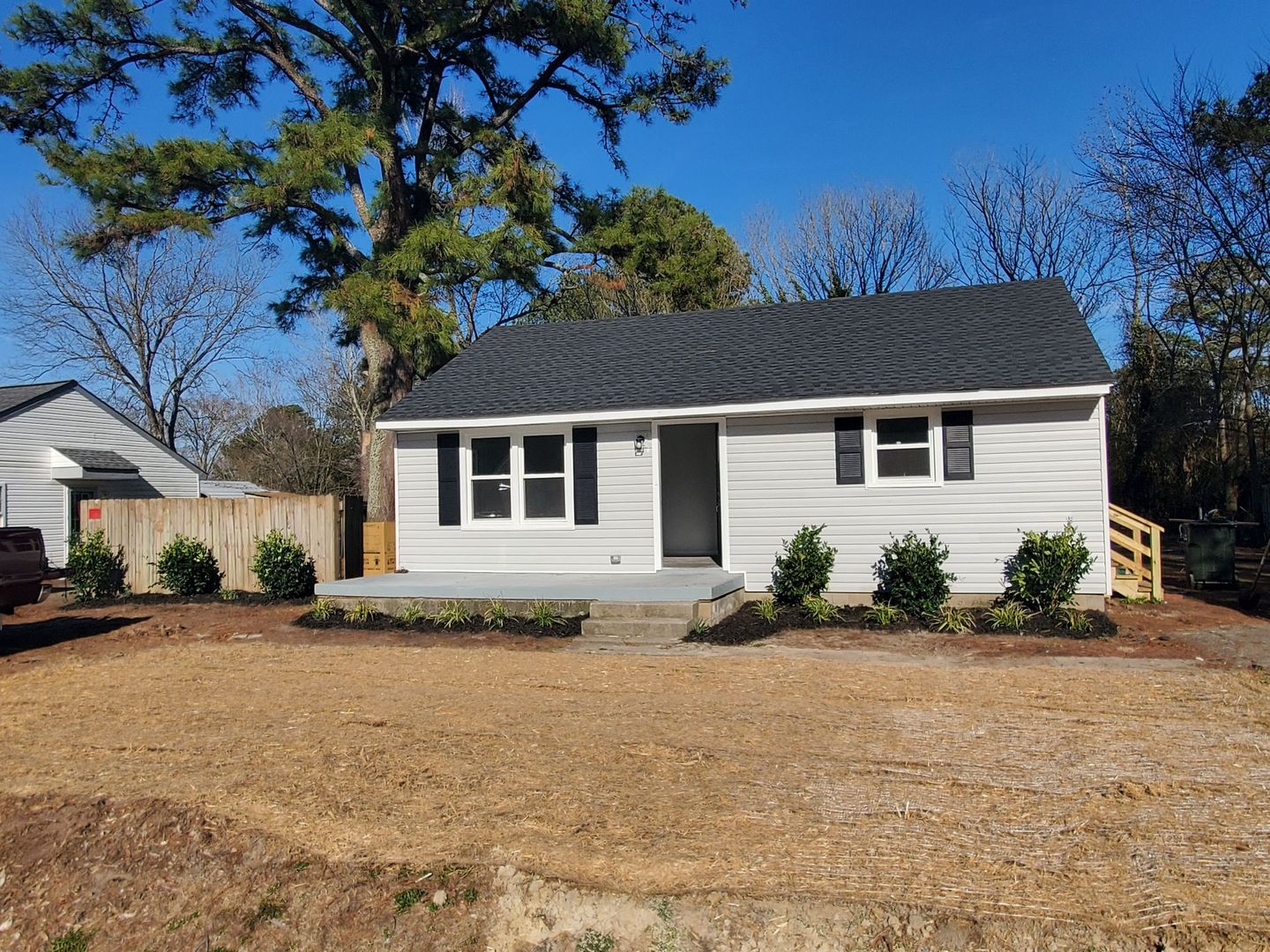 A small, light gray house with a dark roof and black shutters on a sunny day. Landscaping in front.