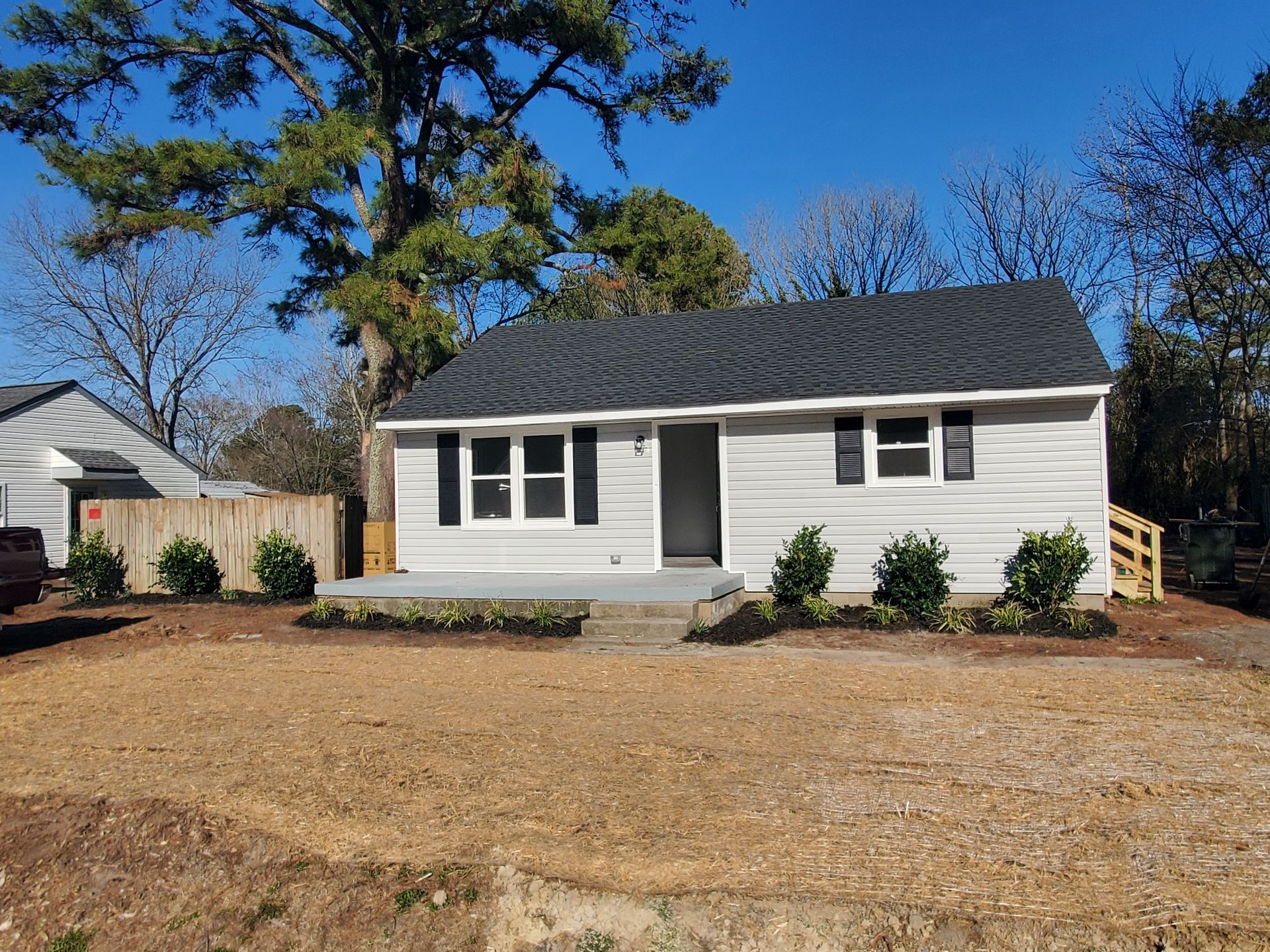 A small, light gray house with a dark roof and black shutters on a sunny day. Landscaping in front.