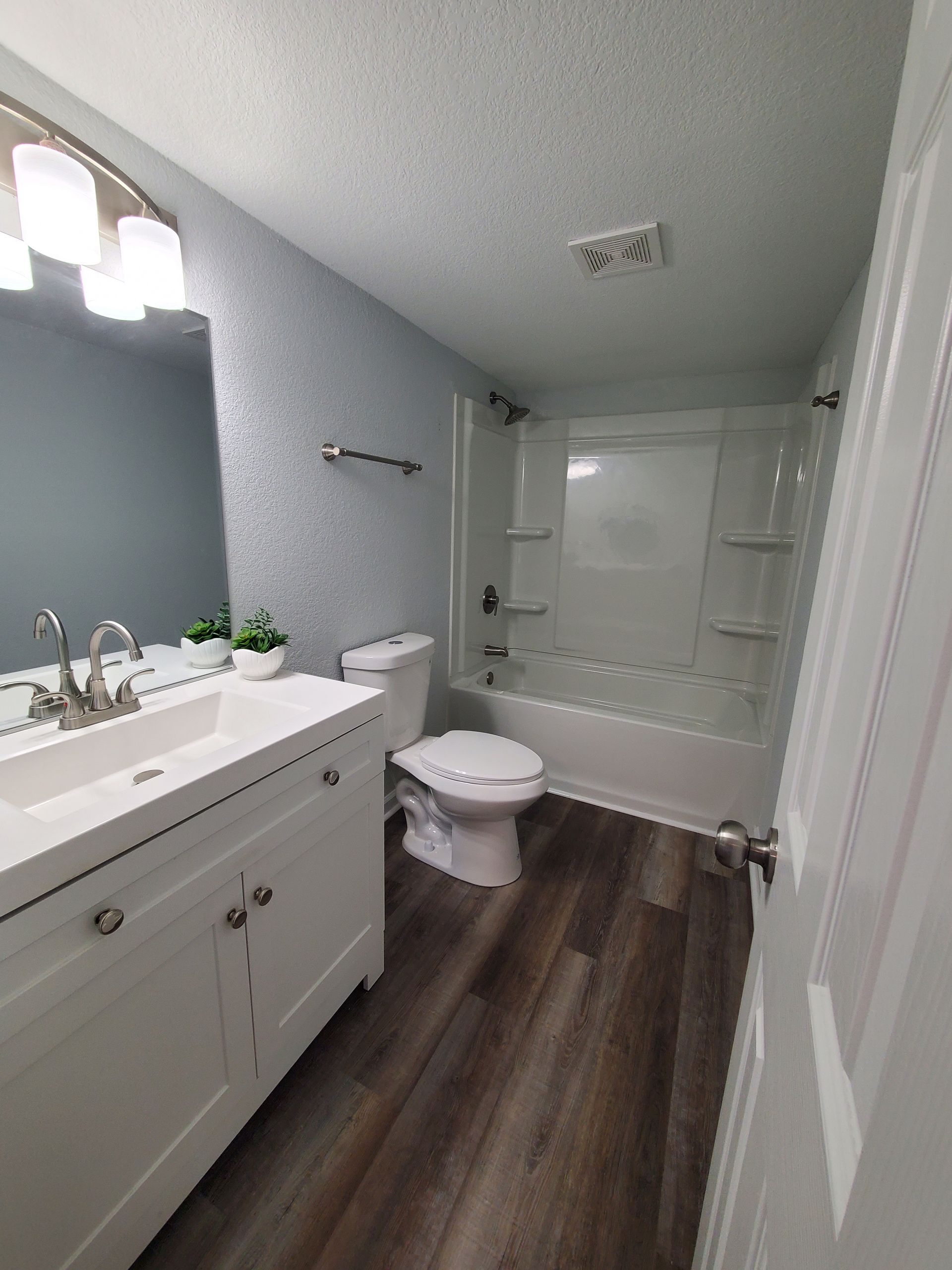 A small bathroom with blue walls, white fixtures, and dark wood-look flooring. 