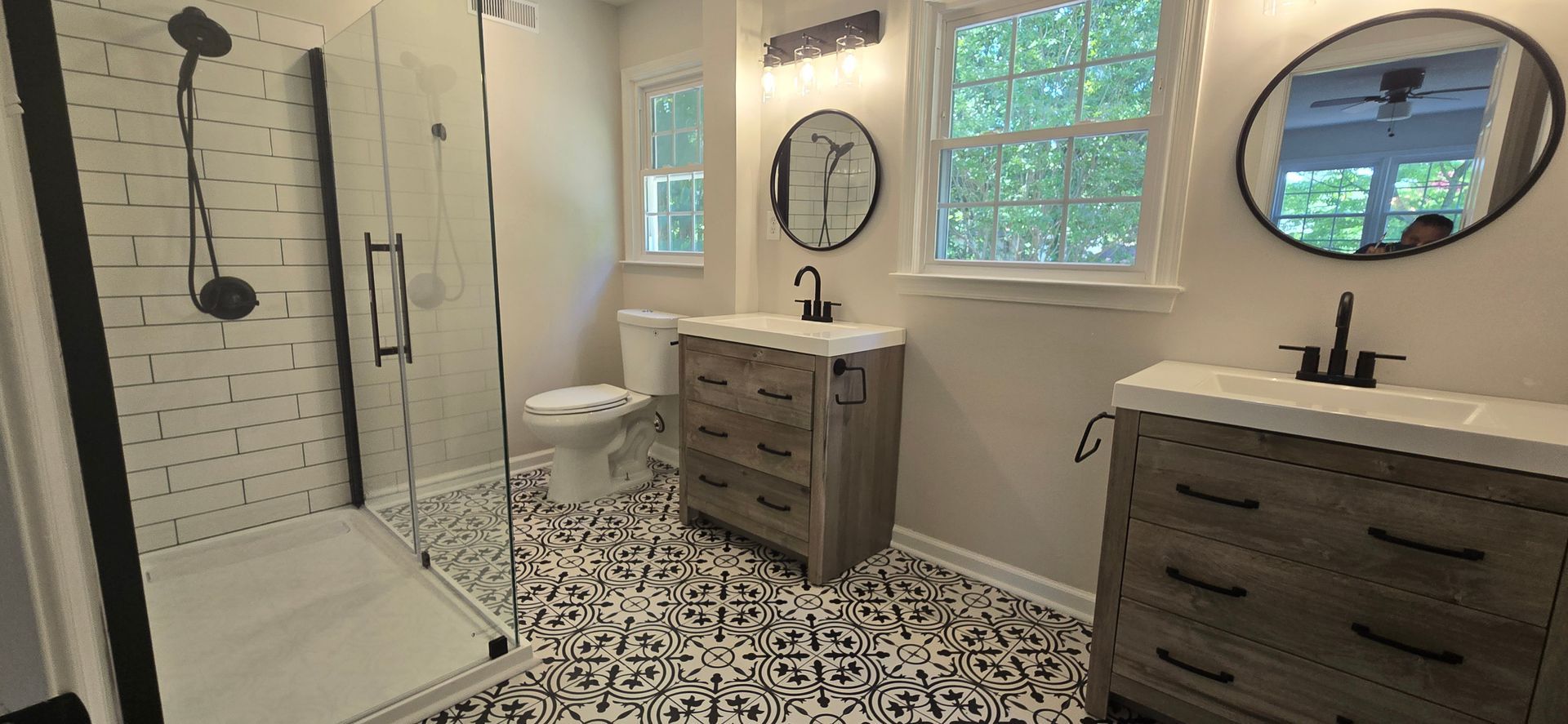 Bathroom with patterned tile floor, two vanities with round mirrors, a shower with black trim, and a toilet.