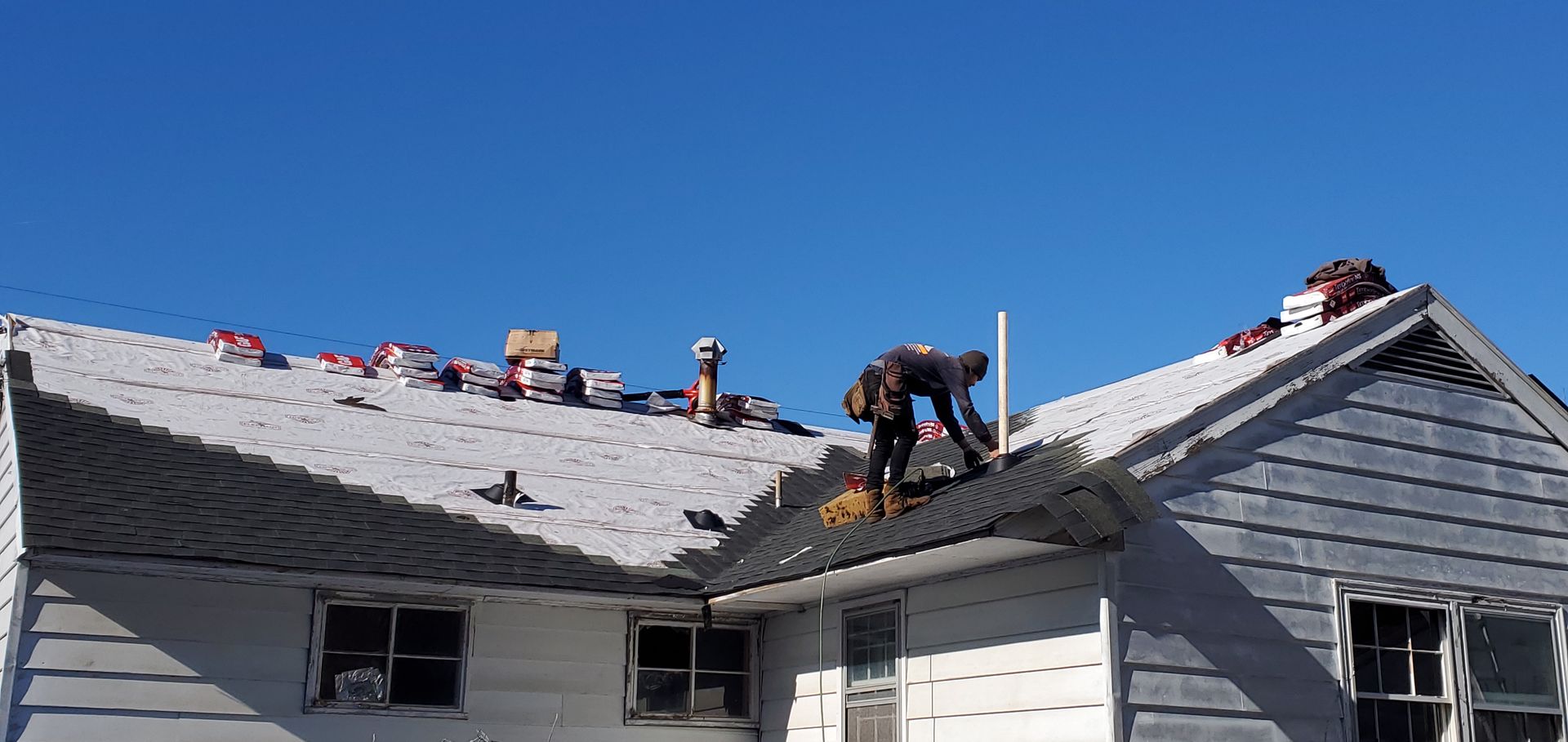 A person working on a roof, surrounded by roofing materials, with a bright blue sky overhead. 