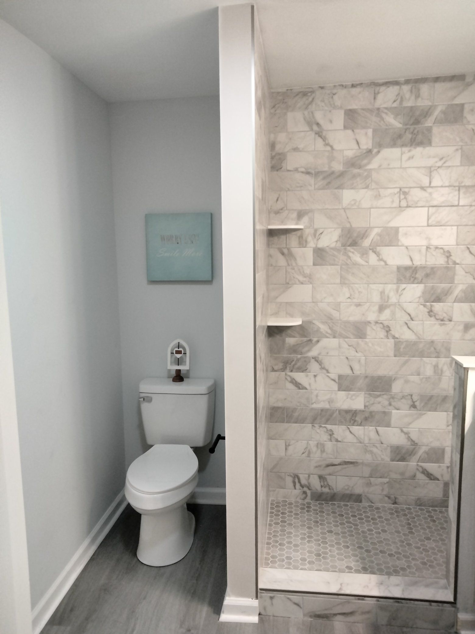 A small bathroom with a white toilet, grey-tiled wall, and a light blue artwork. The floor is a light grey wood-look.