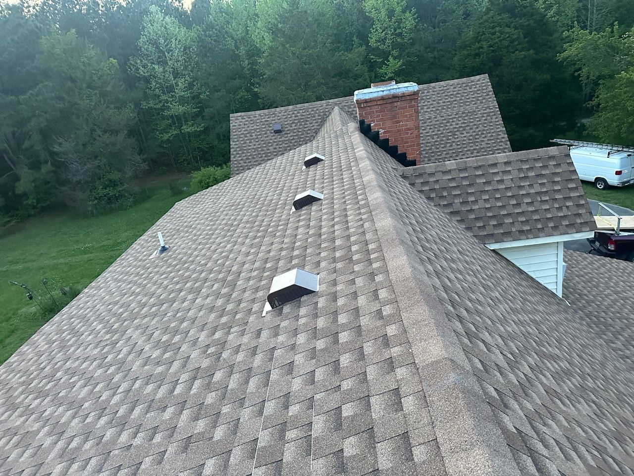 A brown shingled roof with a brick chimney and three vents, set against a backdrop of green trees and a blue sky.