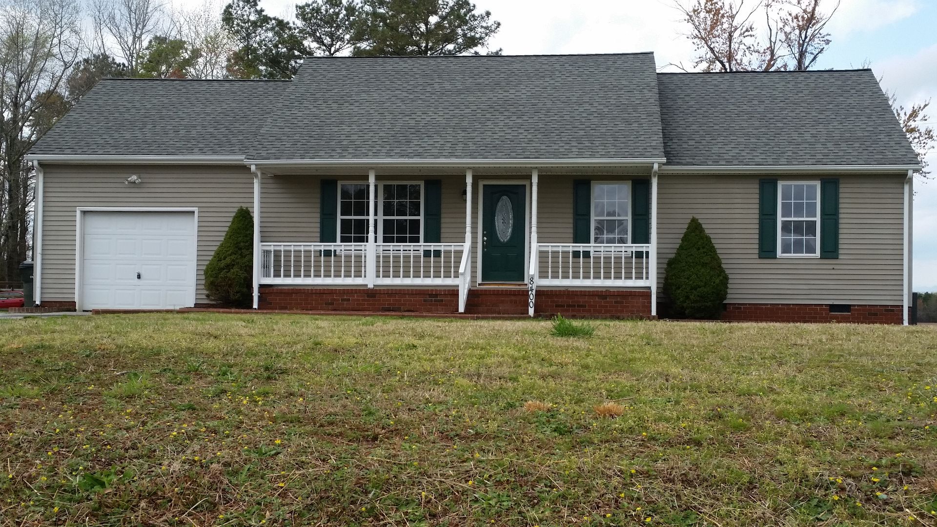 Beige ranch house with a small porch, green shutters, and attached garage. The house sits on a grassy lawn.