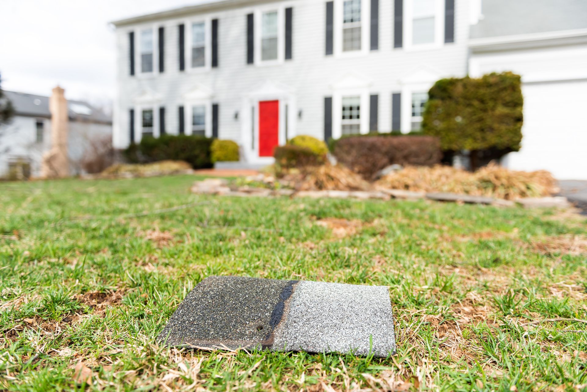 A gray, rectangular, textured tile in a grassy yard in front of a two-story white house with a red door.