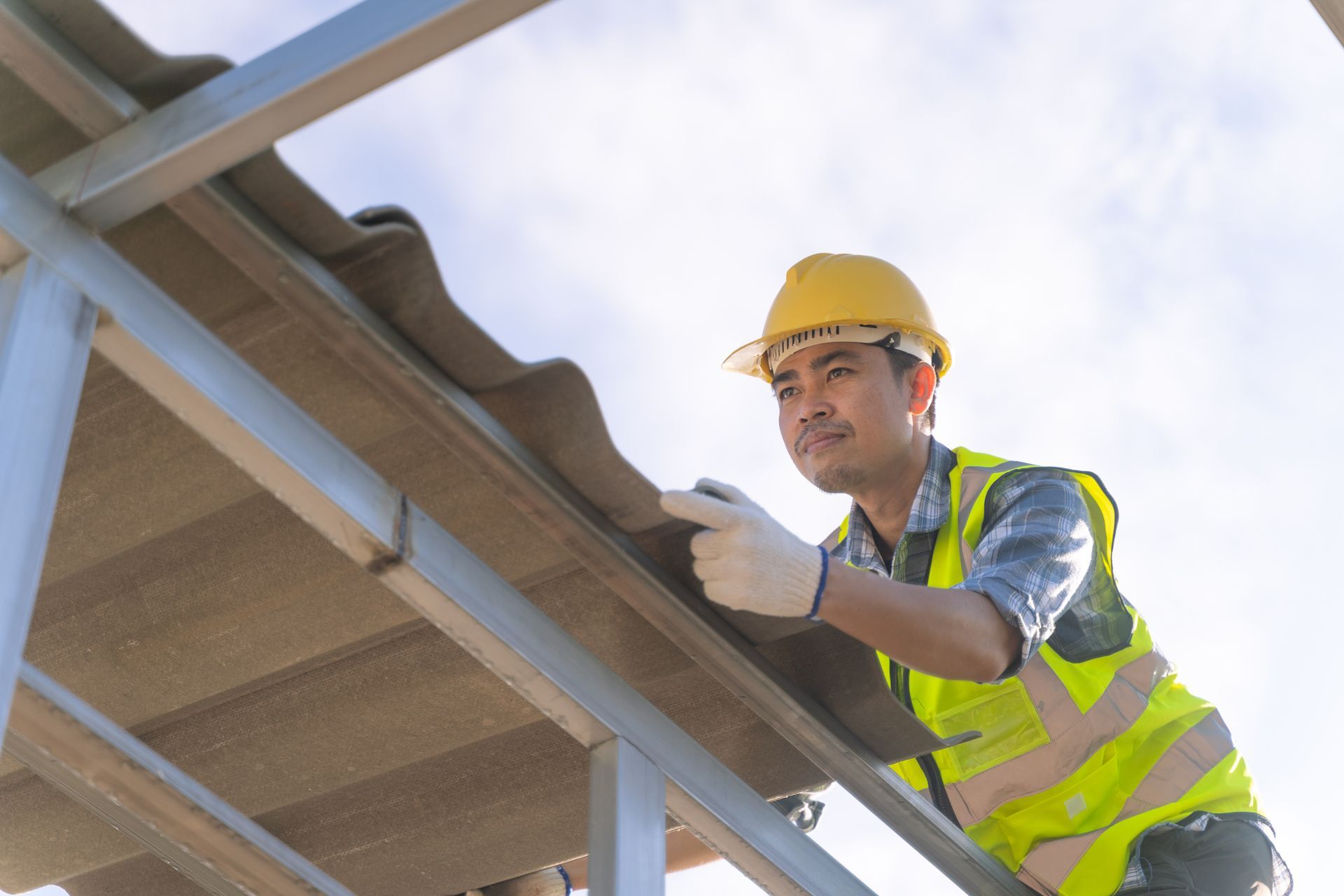 A male Asian roofer in safety gear installs a roof panel on a metal structure under a clear sky