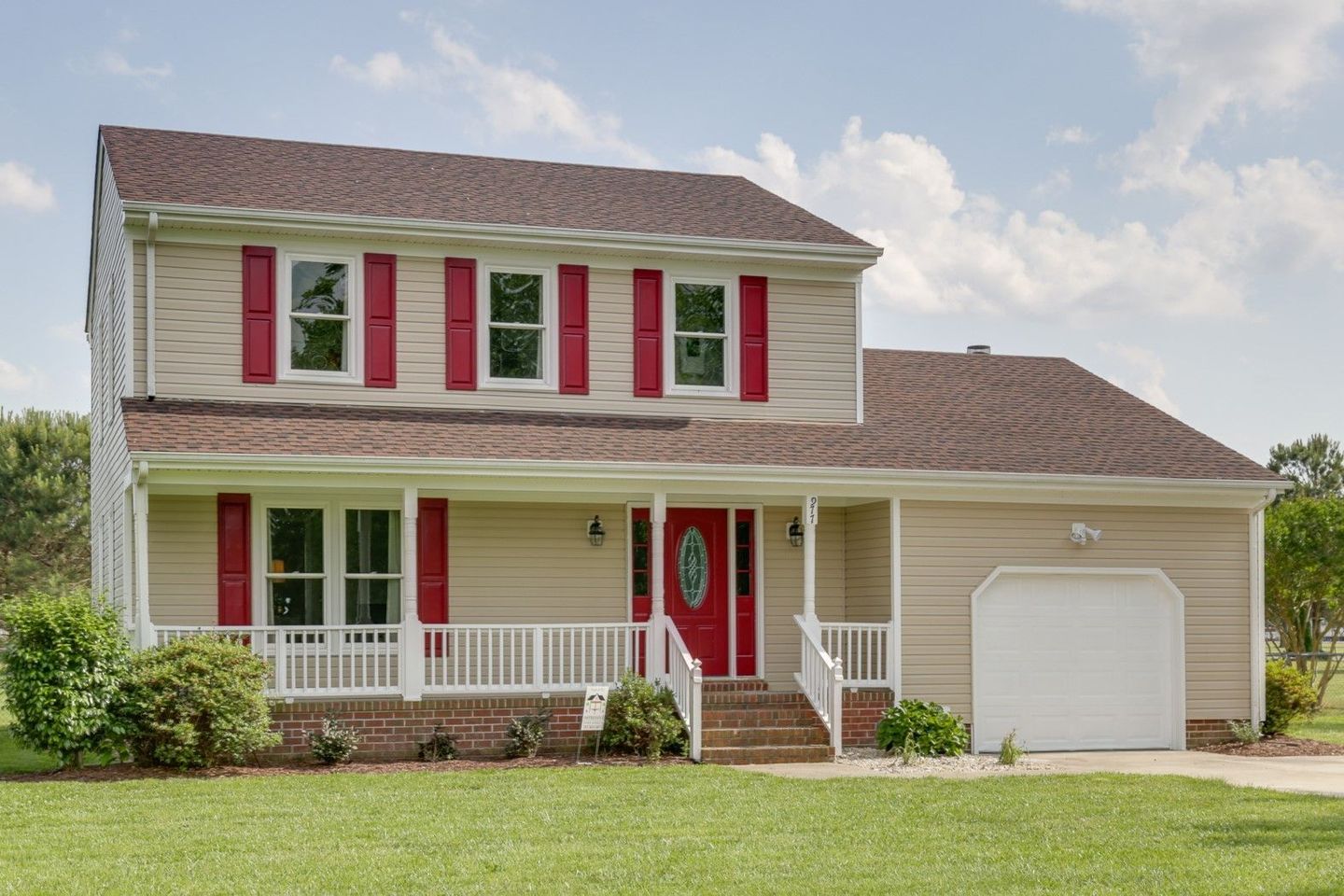 Beige two-story house with red shutters and door, white porch, and attached garage.
